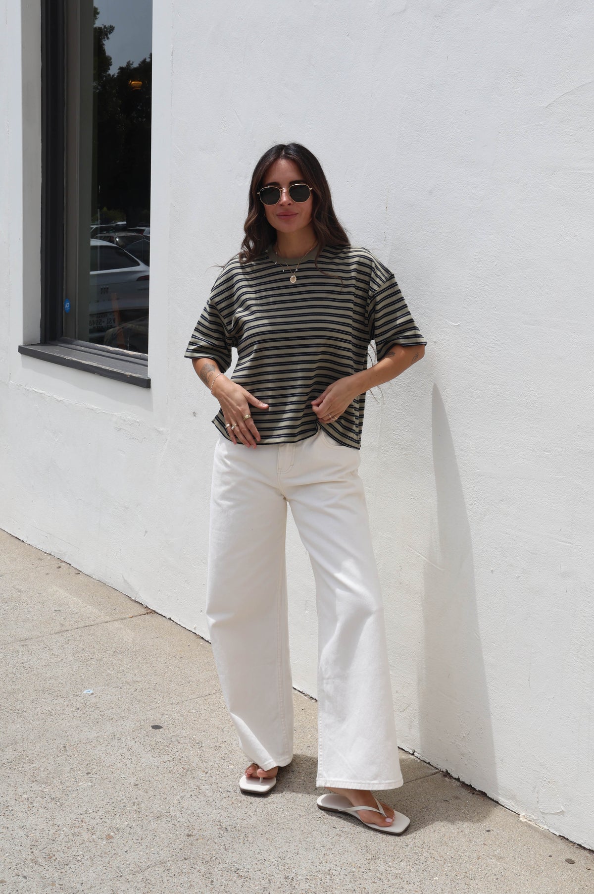 A woman stands outdoors against a white stucco wall wearing a black and white horizontally striped short-sleeve top, white wide-leg pants, and white slide sandals.