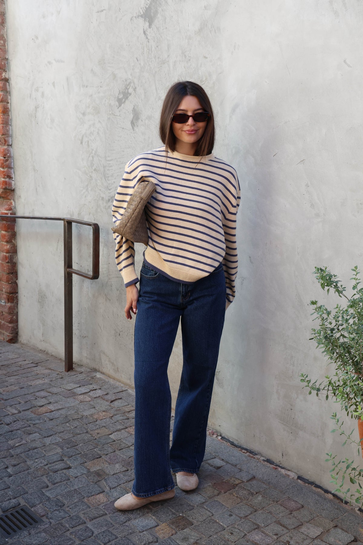 Woman wearing a cream and blue striped sweater with jeans and sunglasses standing against a plain background.