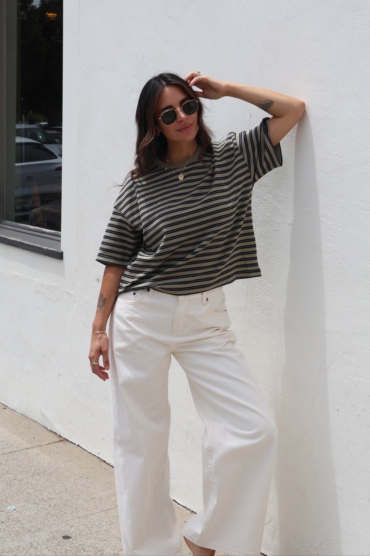 A woman wearing a dark green and black horizontally striped short-sleeve t-shirt and white wide-leg pants, leaning against a white stucco wall outdoors.