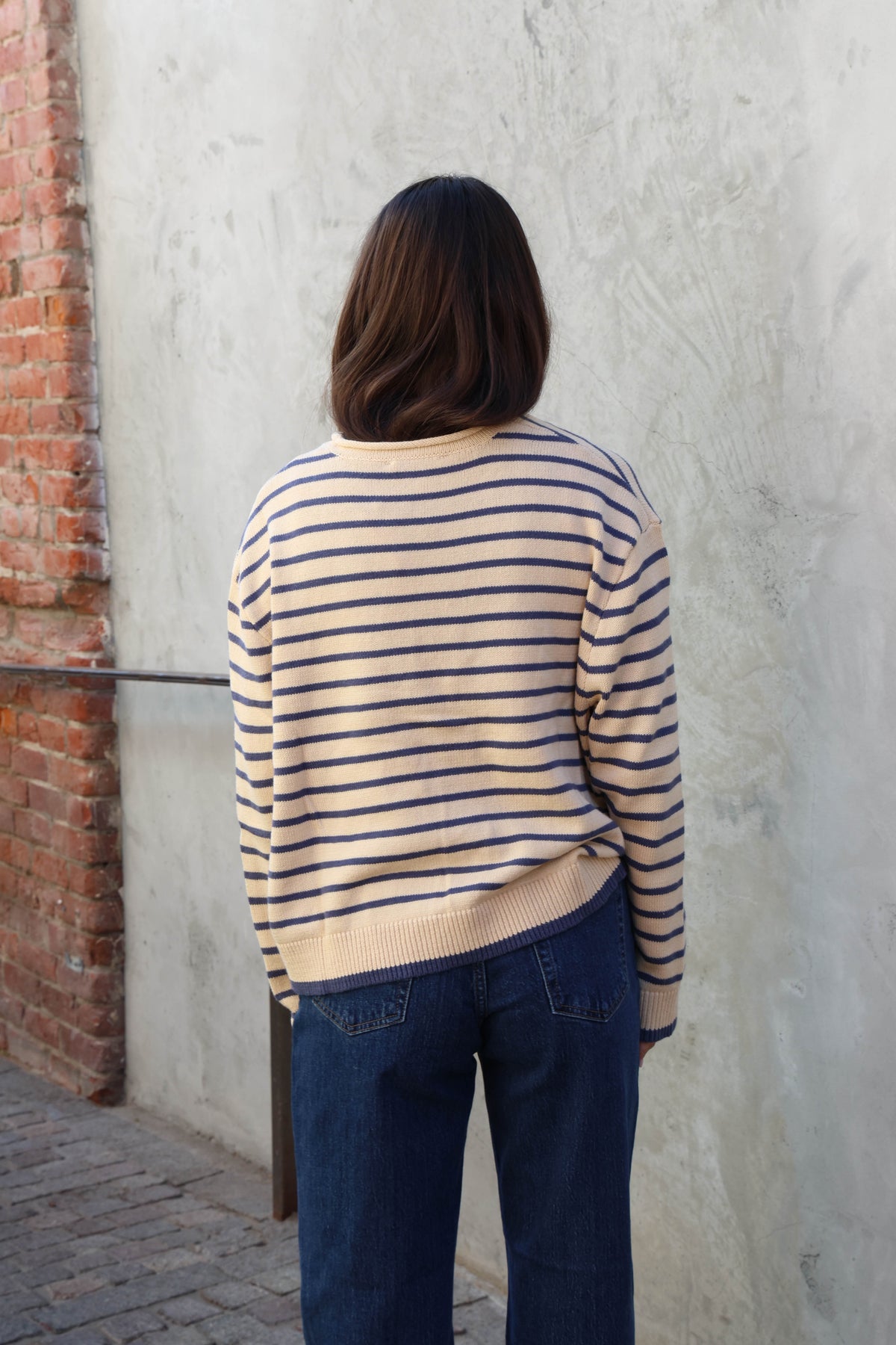 Woman wearing a cream and blue striped sweater with jeans and sunglasses standing against a plain background.