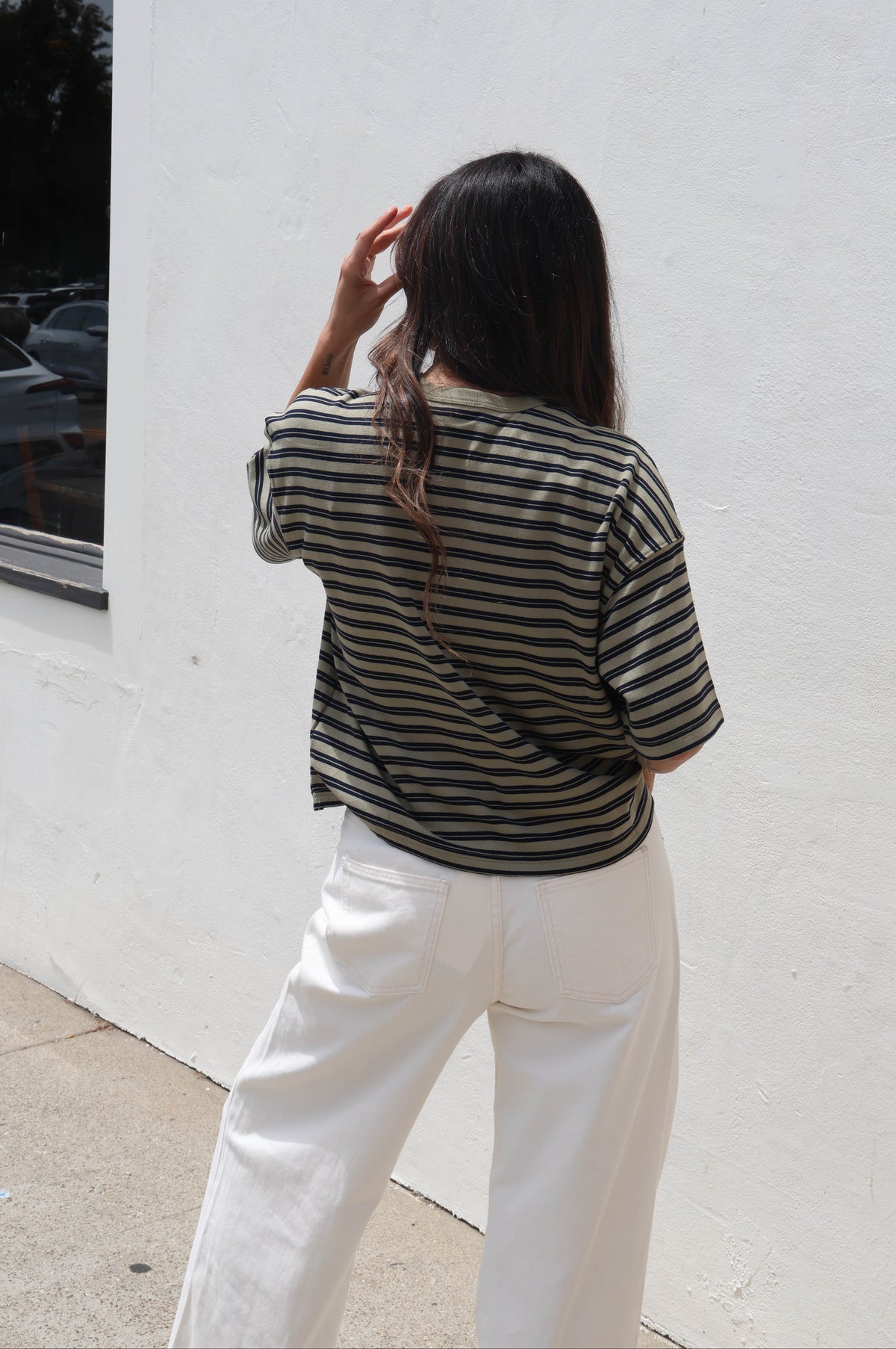 A woman viewed from behind wearing a black and white horizontally striped short-sleeve top and white wide-leg pants, standing outdoors against a white stucco wall.