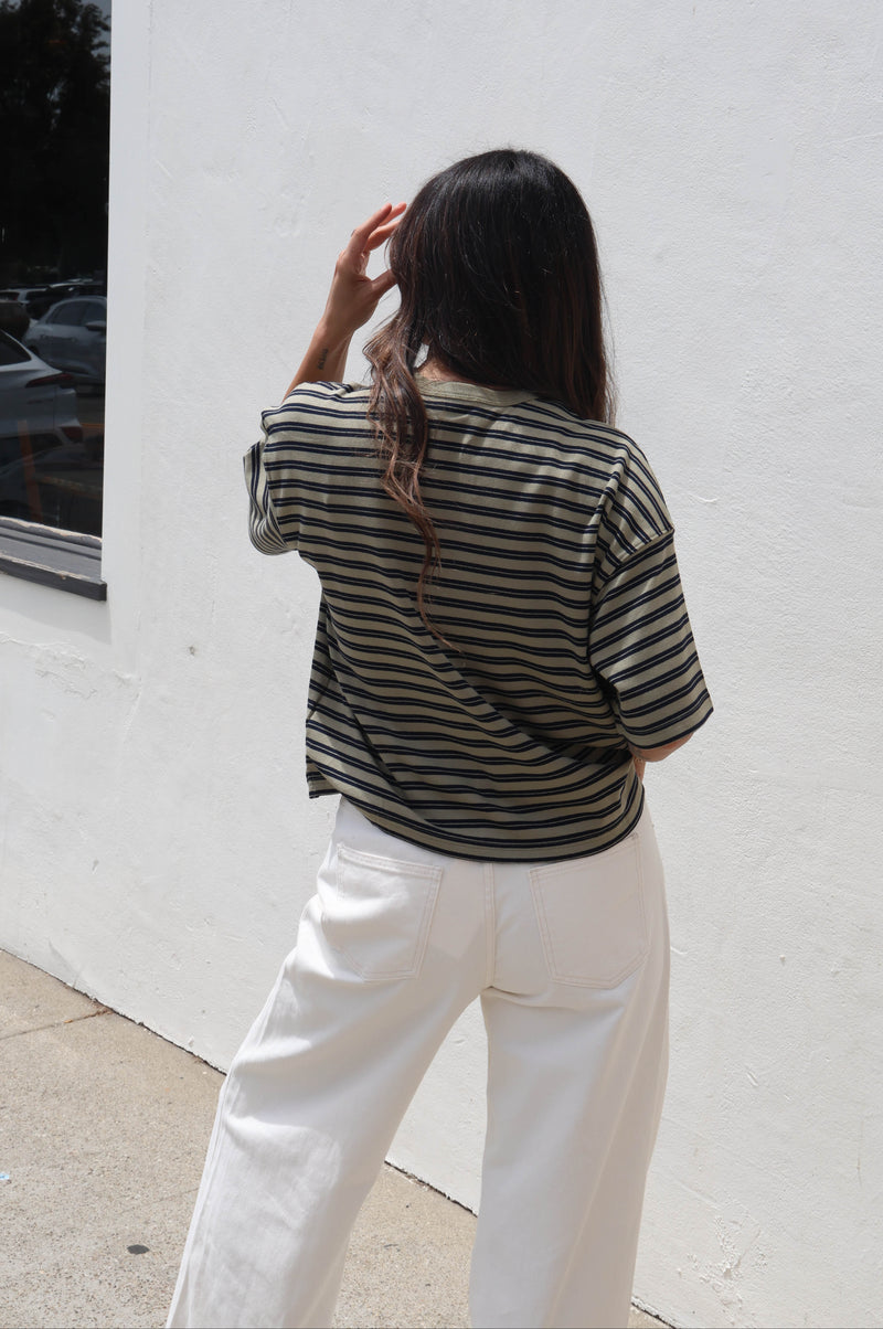 A woman viewed from behind wearing a black and white horizontally striped short-sleeve top and white wide-leg pants, standing outdoors against a white stucco wall.