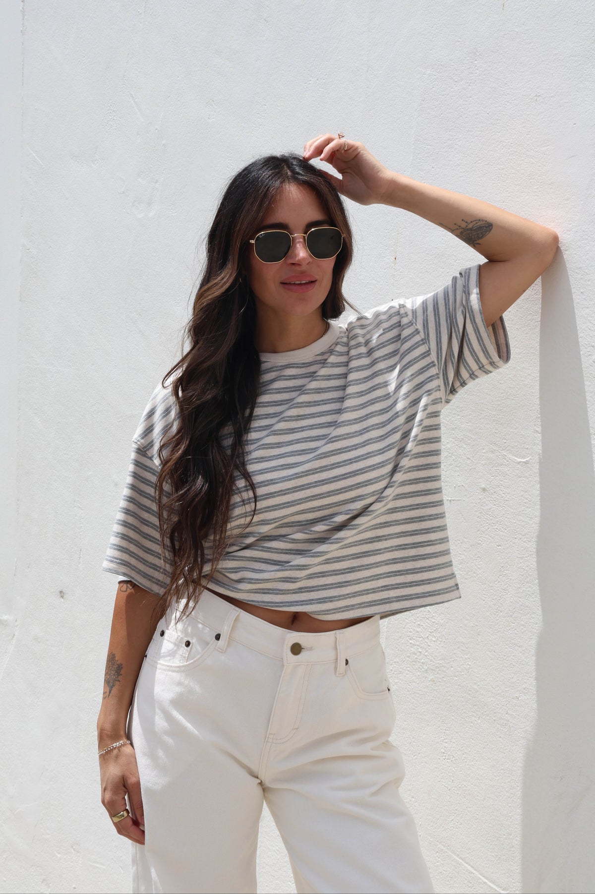 A woman wearing a gray and white horizontally striped short-sleeve crop top and white pants, posing against a white stucco wall.