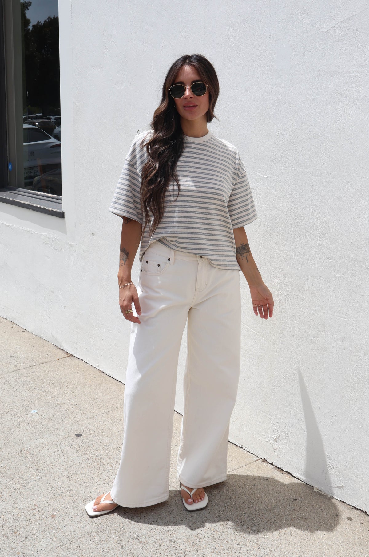 A woman stands outdoors against a white stucco wall wearing a gray and white striped short-sleeve t-shirt, white wide-leg pants, and white sandals.