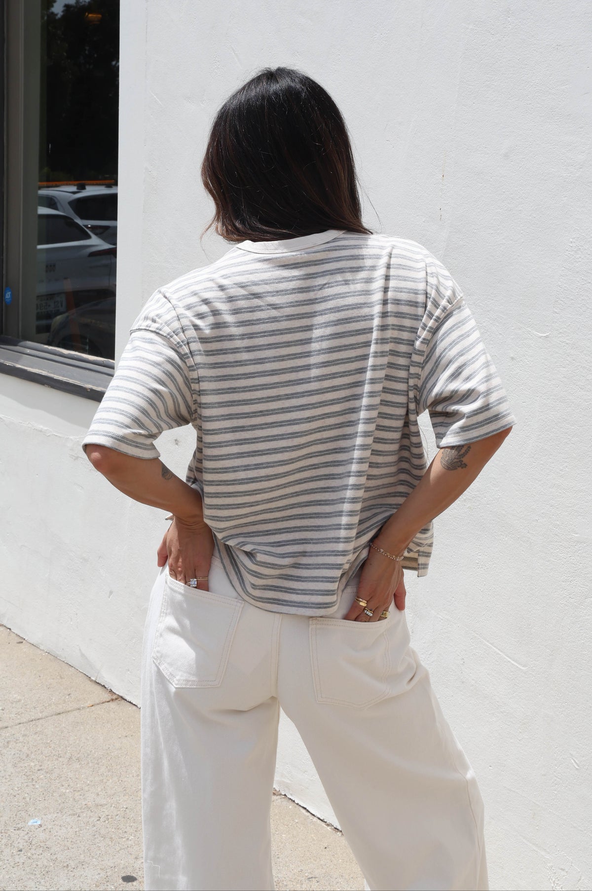 A person viewed from behind wearing a gray and white horizontally striped short-sleeve t-shirt and white pants, standing outdoors against a white stucco wall.