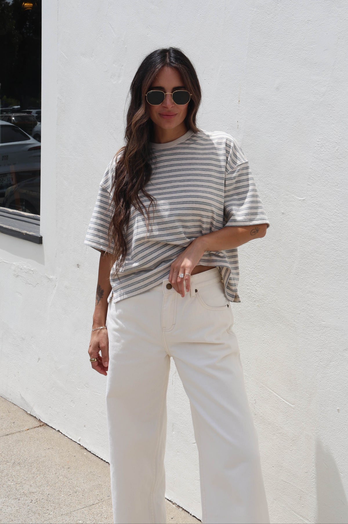 A woman wearing a gray and white horizontally striped t-shirt and white wide-leg pants, standing outdoors against a white stucco wall.