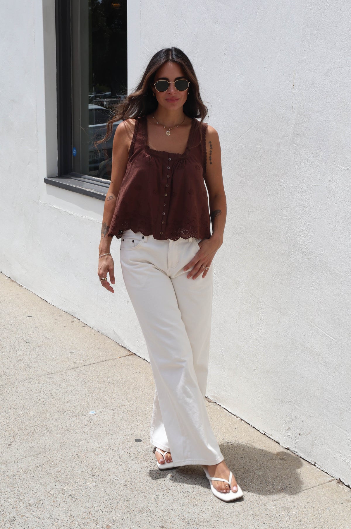 A woman wearing a dark brown sleeveless top with a scalloped edge and white pants, standing outdoors against a white stucco wall.