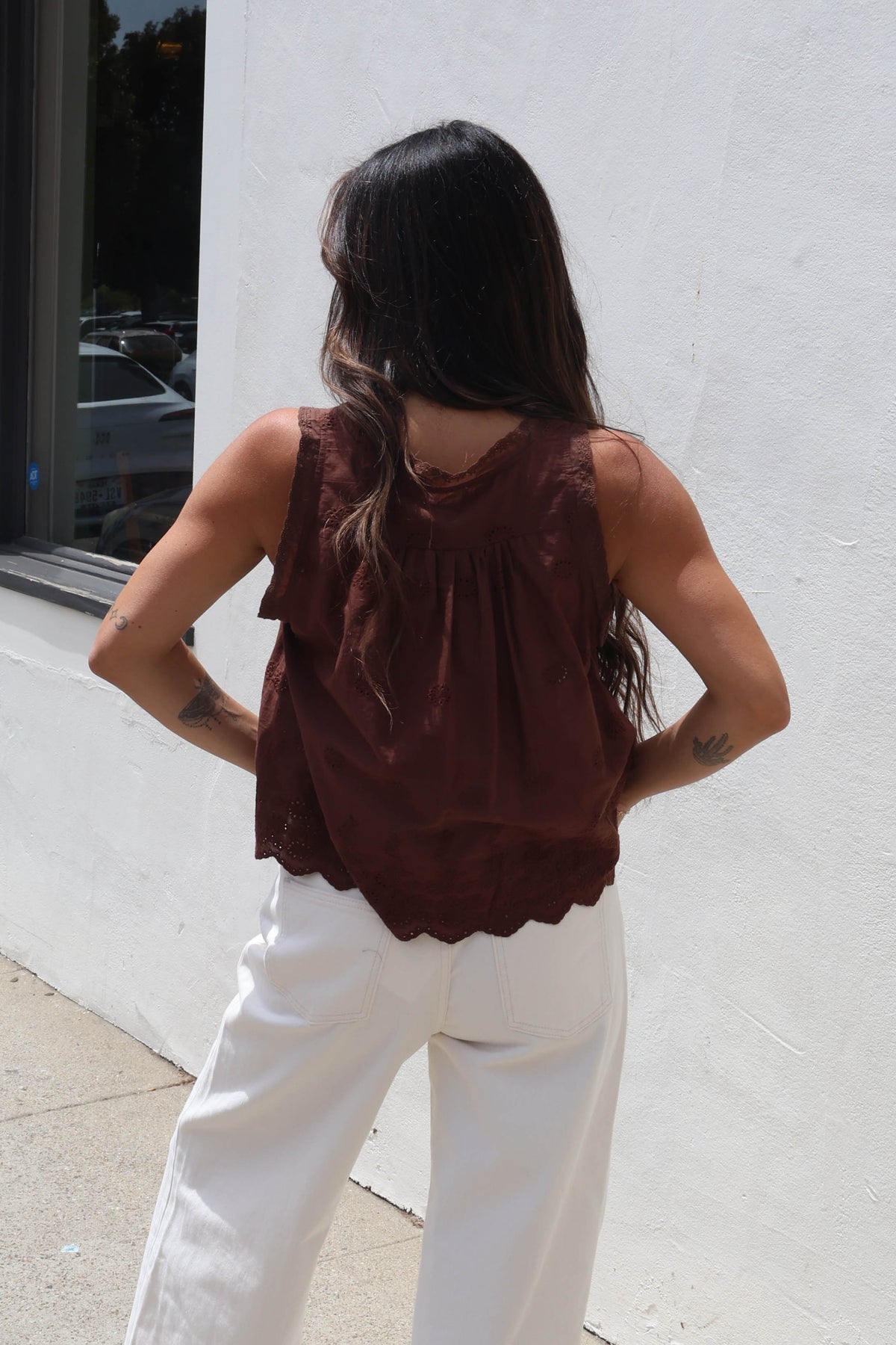 A woman shown from behind wearing a sleeveless dark brown ruffled top and white pants, standing outdoors against a white wall.