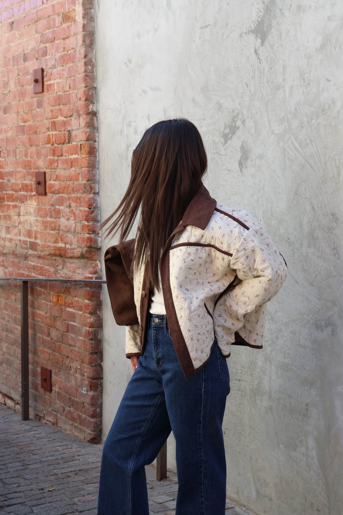 Woman wearing a off white jacket, brown pants holding a brown bag in front of a plain wall.