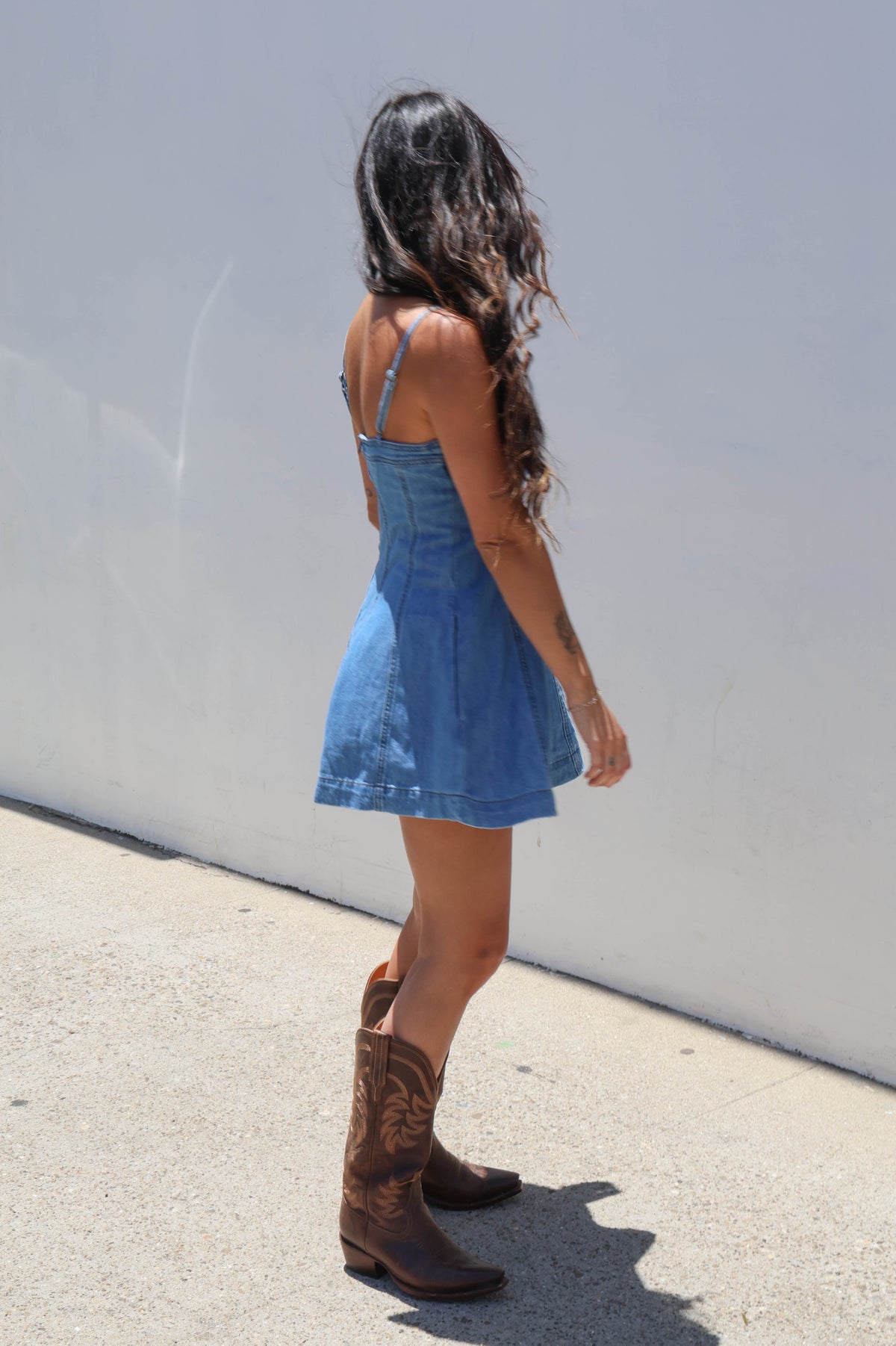 Woman wearing a denim dress and cowboy boots against a white wall.