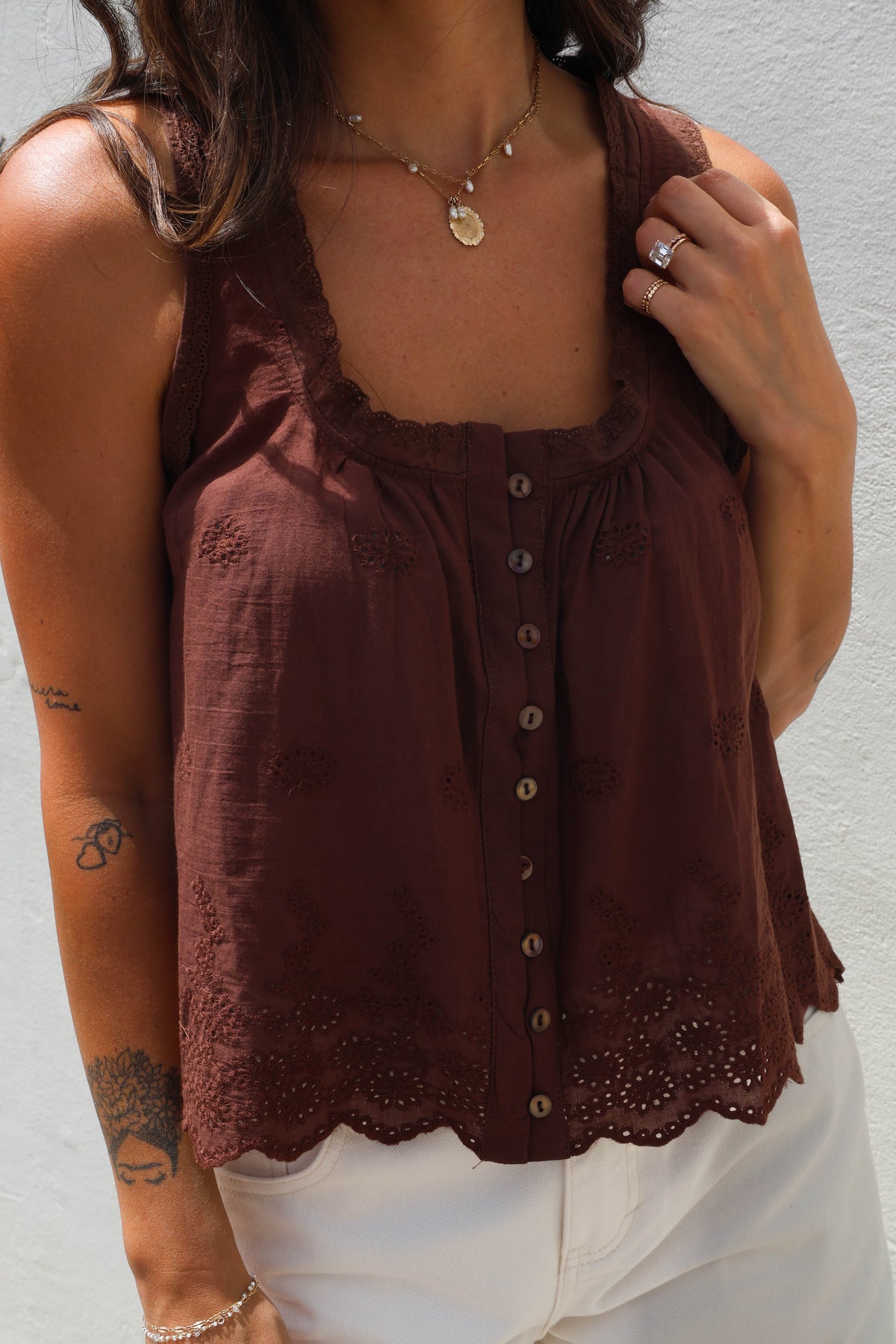 A woman wearing a sleeveless brown top with a scalloped eyelet hem and front buttons, paired with white pants, photographed against a light wall.