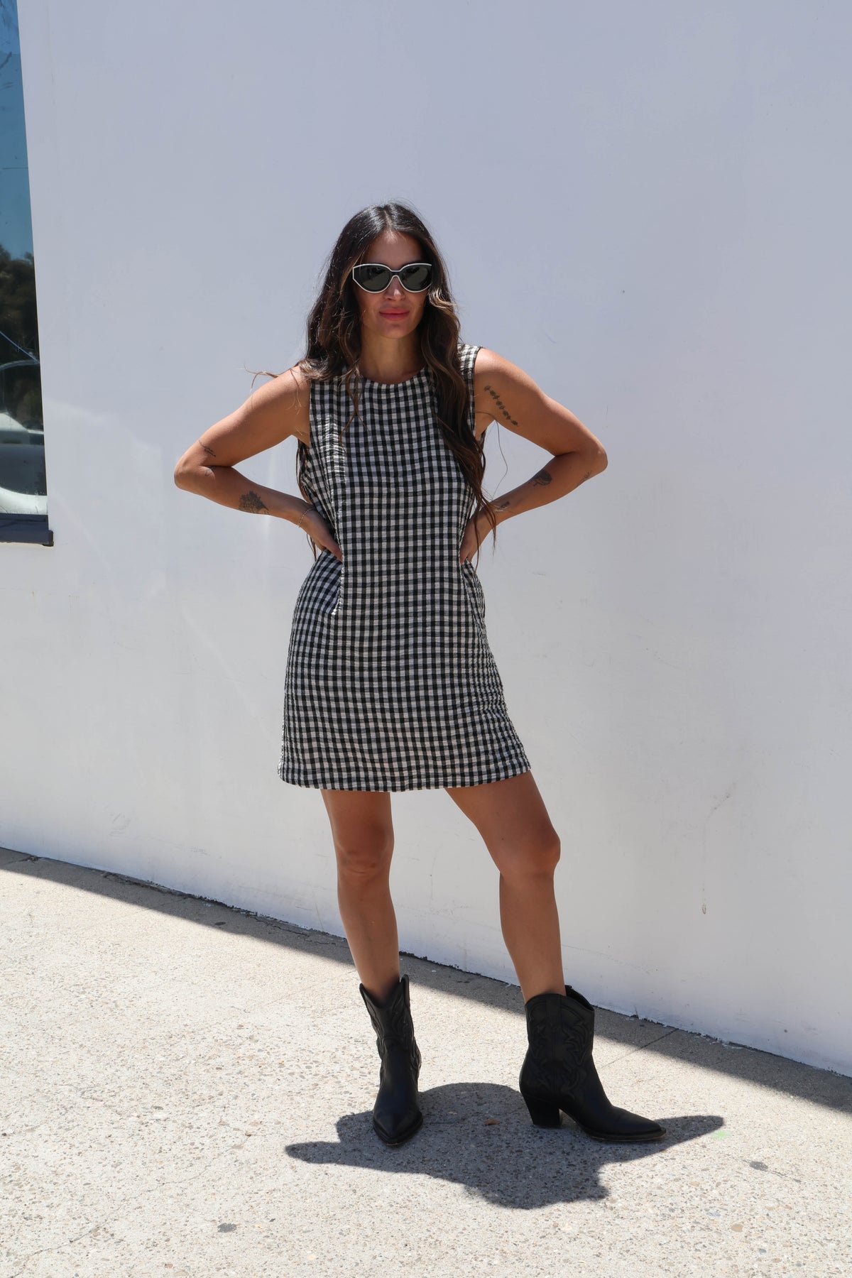 Woman in a checkered dress and black boots standing against a white wall.