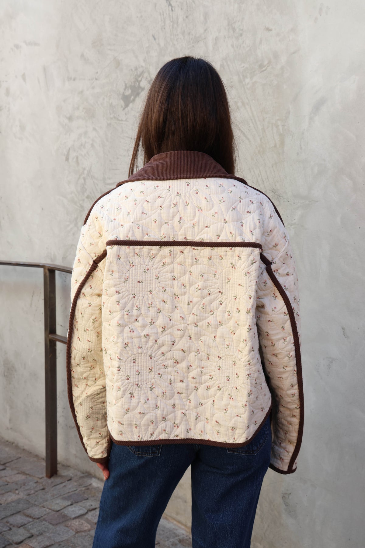 Woman wearing a brown jacket, brown pants holding a brown bad in front of a plain wall.