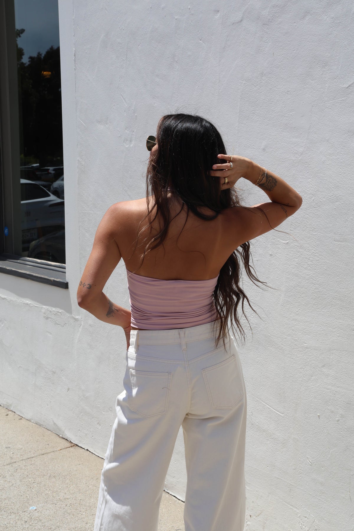 A woman wearing a light blush halter top and white jeans, standing outdoors against a white wall.