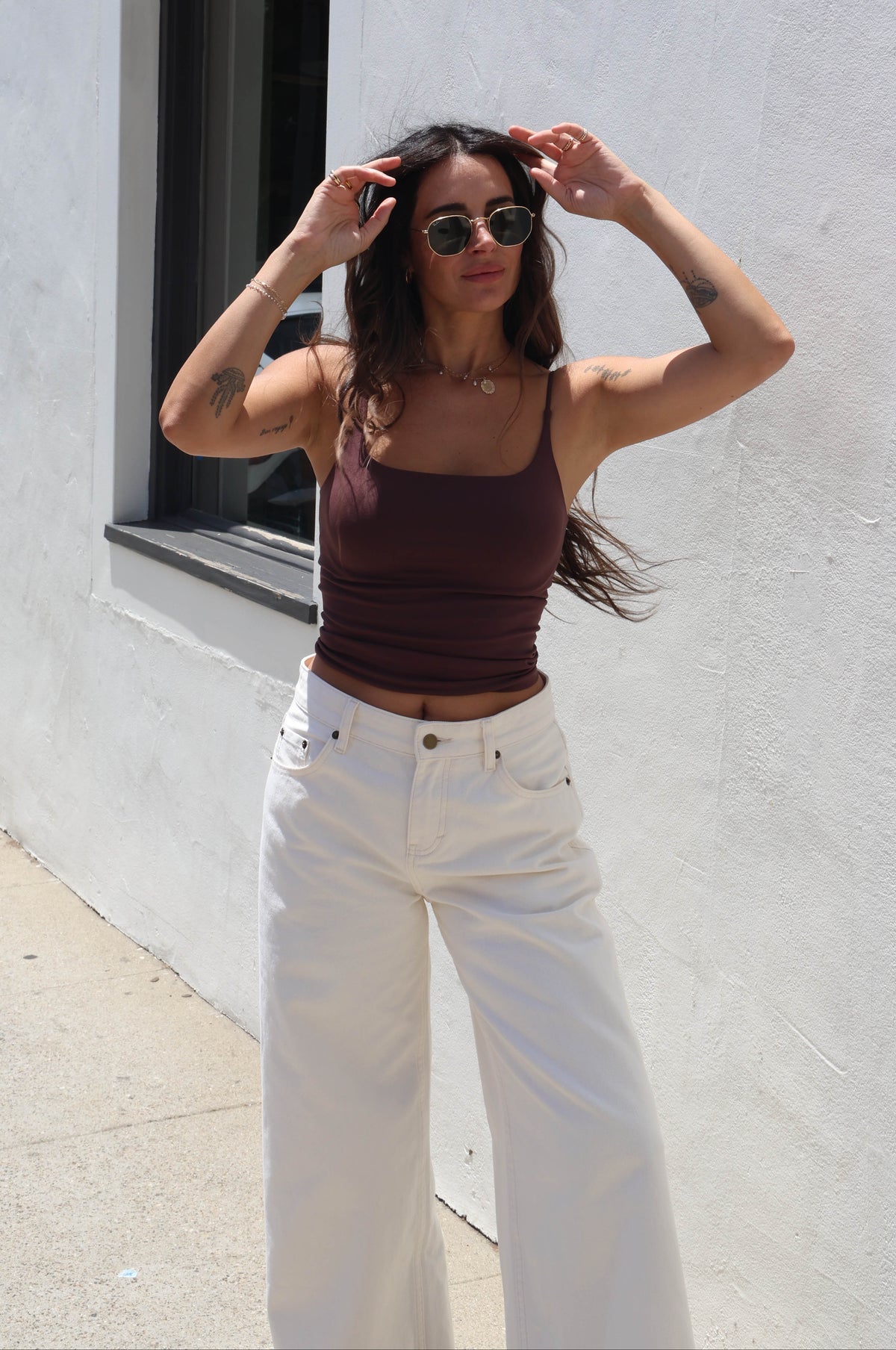 A woman posing outdoors against a white stucco wall, wearing a dark brown ribbed tank top and white wide-leg jeans.