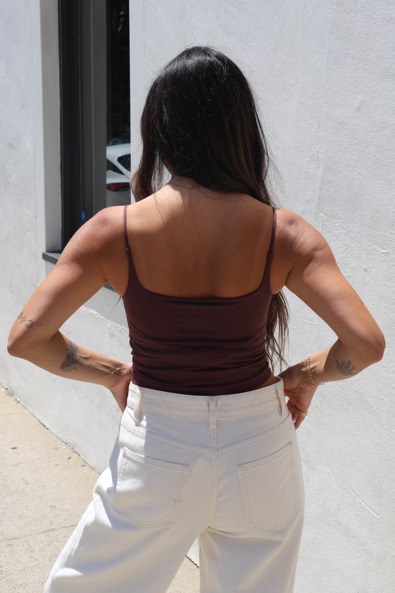 A woman shown from behind wearing a dark brown spaghetti-strap camisole top and white pants, standing outdoors against a white stucco wall.