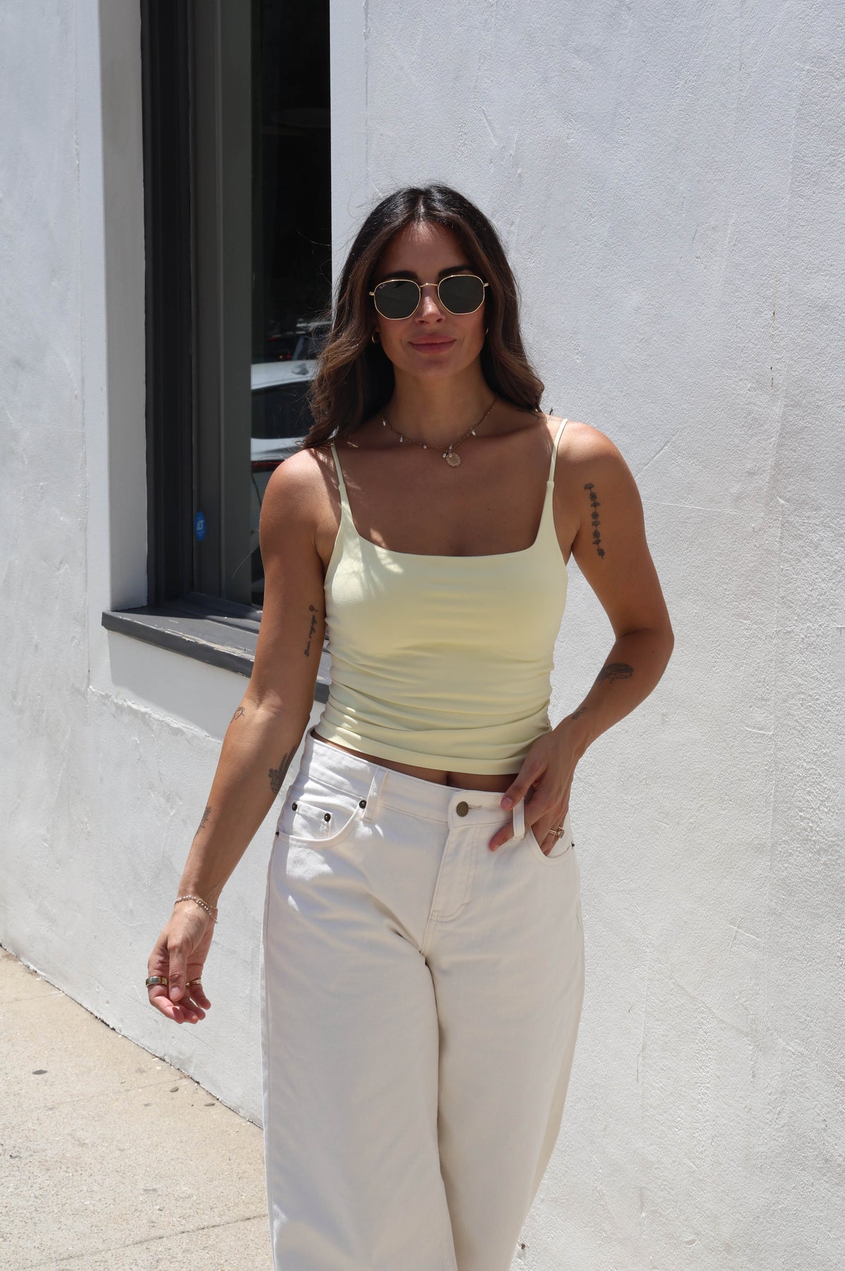 A woman wearing a pale yellow ribbed tank top and white jeans stands outdoors against a white stucco wall.