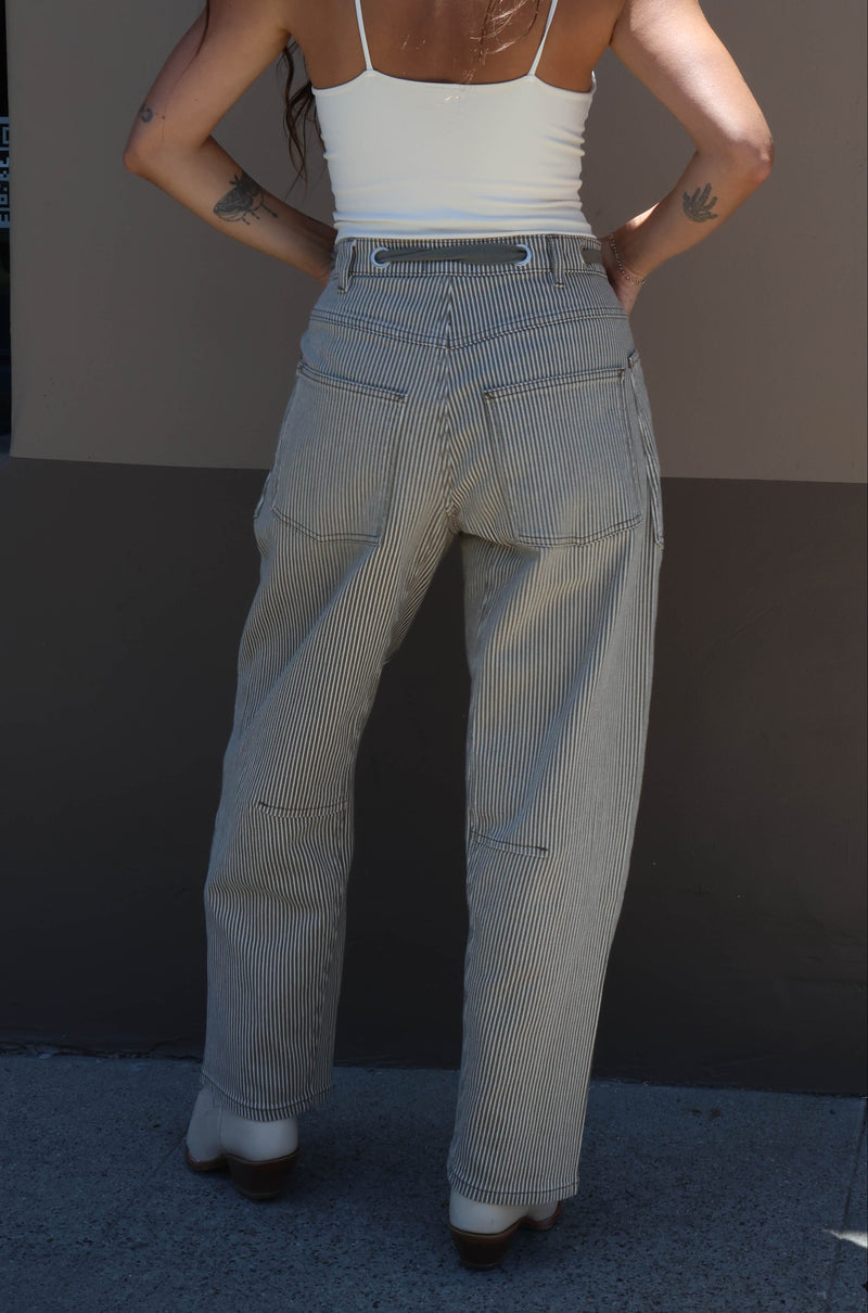 Person wearing a white tank top and beige pants standing against a plain wall.