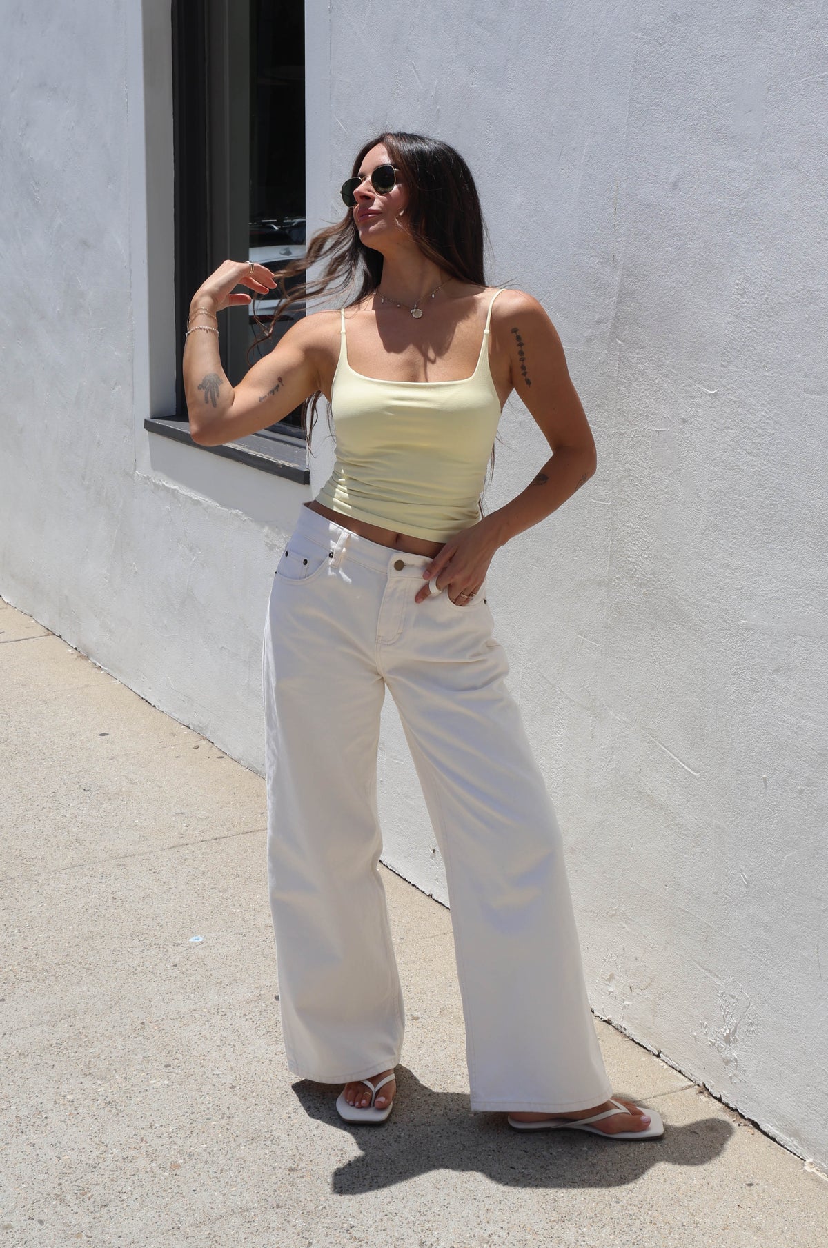 A woman posing outdoors against a white stucco wall, wearing a pale yellow ribbed tank top and white wide-leg jeans with brown sandals.