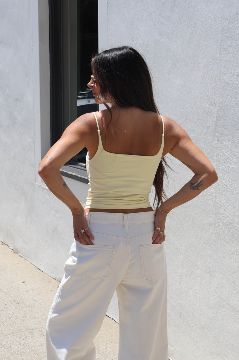 A woman with long dark hair is shown from behind, wearing a pale yellow spaghetti-strap tank top and white wide-leg pants, standing outdoors against a white stucco wall.