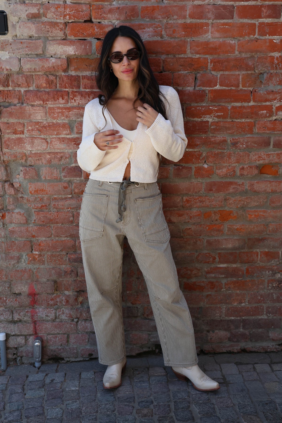 Woman in a white cardigan and beige pants standing against a brick wall.