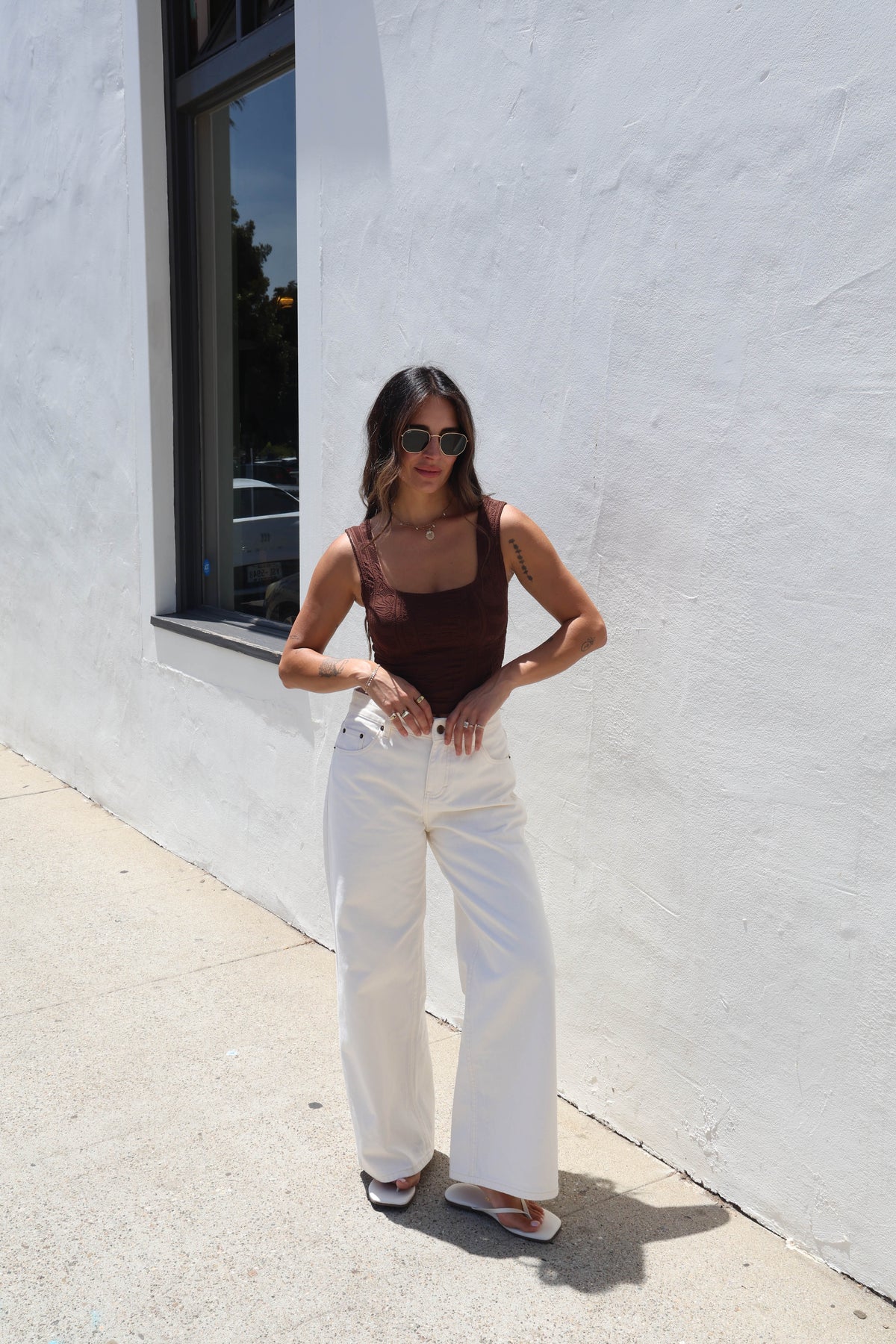 A woman wearing a brown ribbed tank top and white jeans, standing outdoors against a white wall.