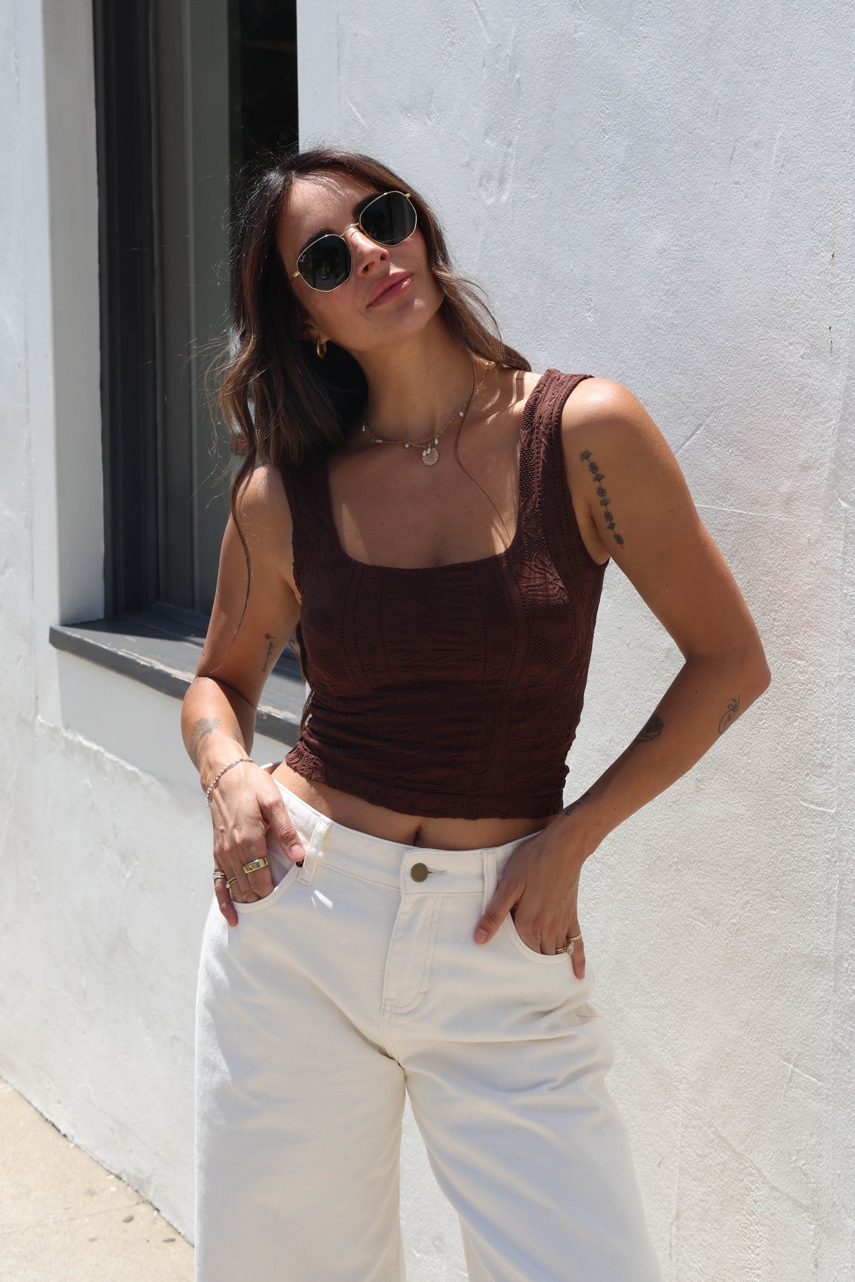 A woman wearing a brown ribbed tank top and white jeans, standing outdoors against a white wall.