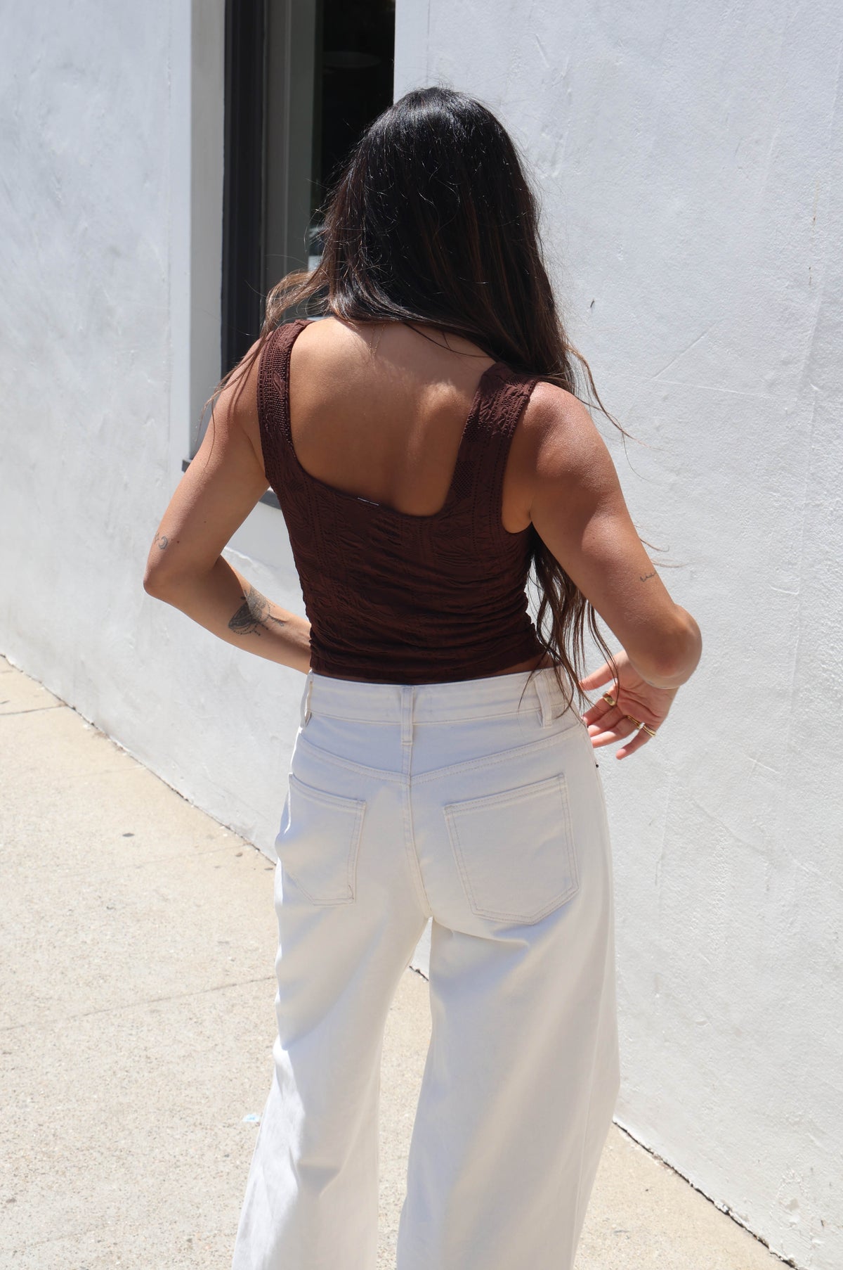 A woman wearing a brown ribbed tank top and white jeans, standing outdoors against a white wall.