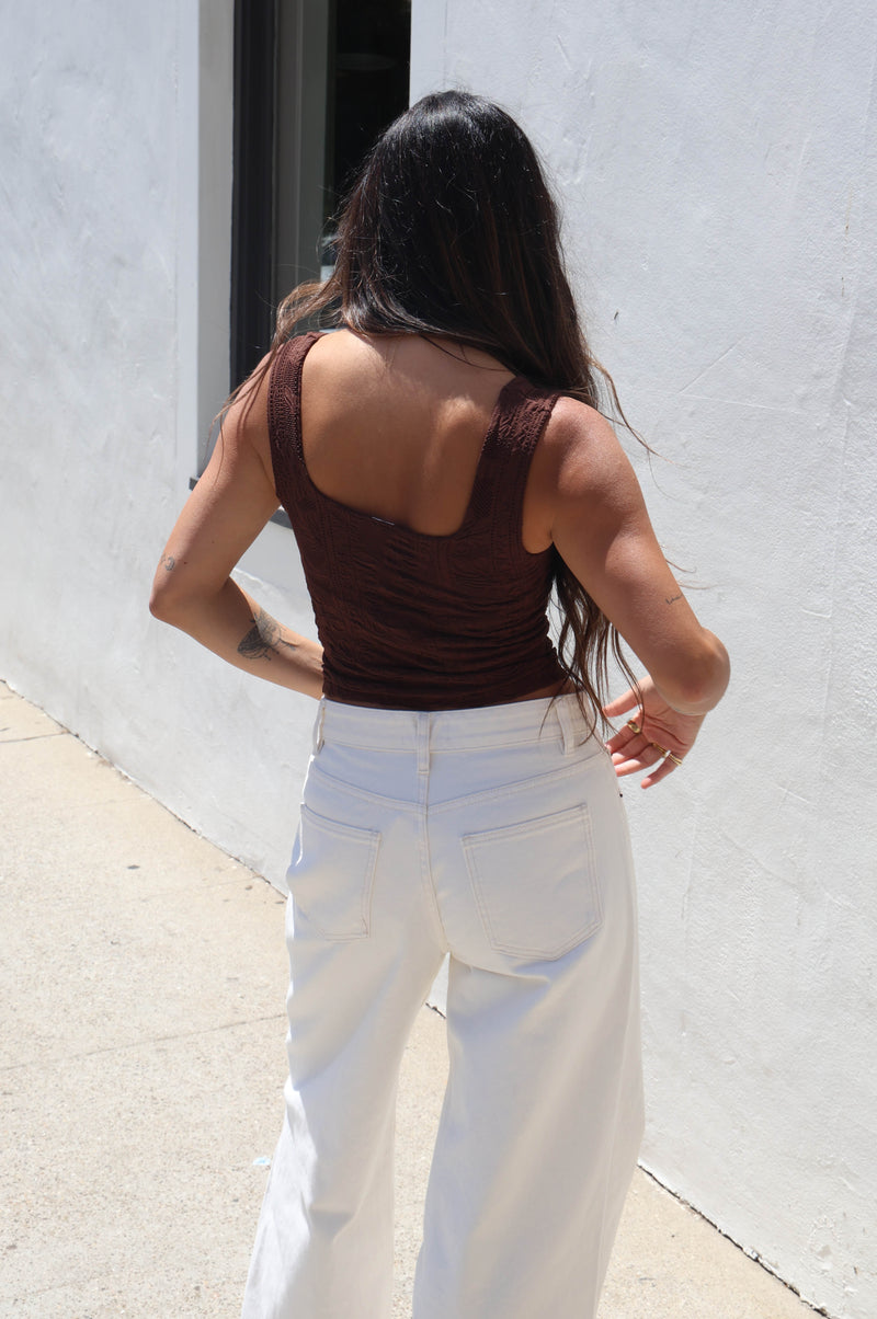 A woman wearing a brown ribbed tank top and white jeans, standing outdoors against a white wall.