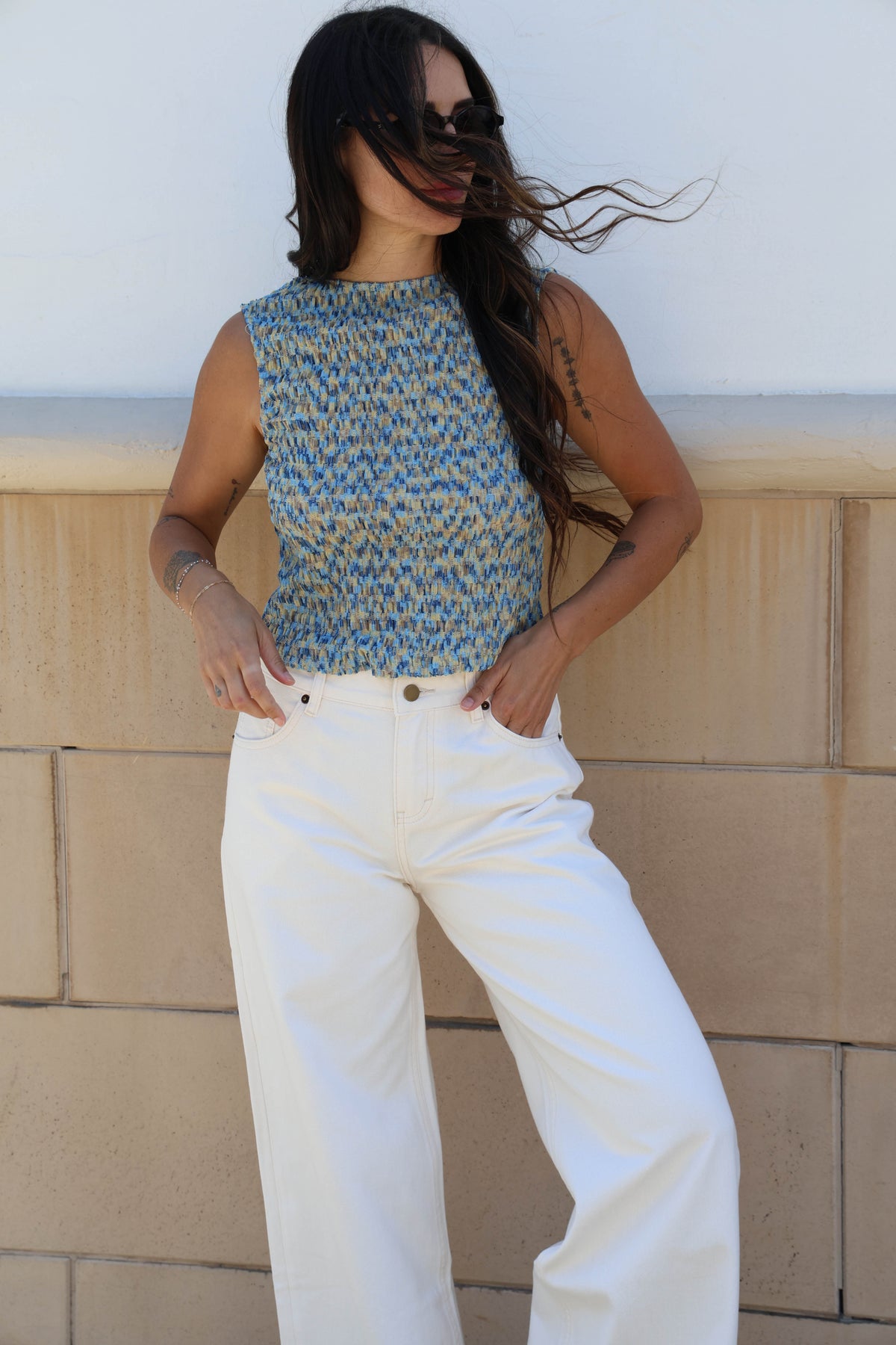 Woman wearing a patterned top and white pants against a beige wall.