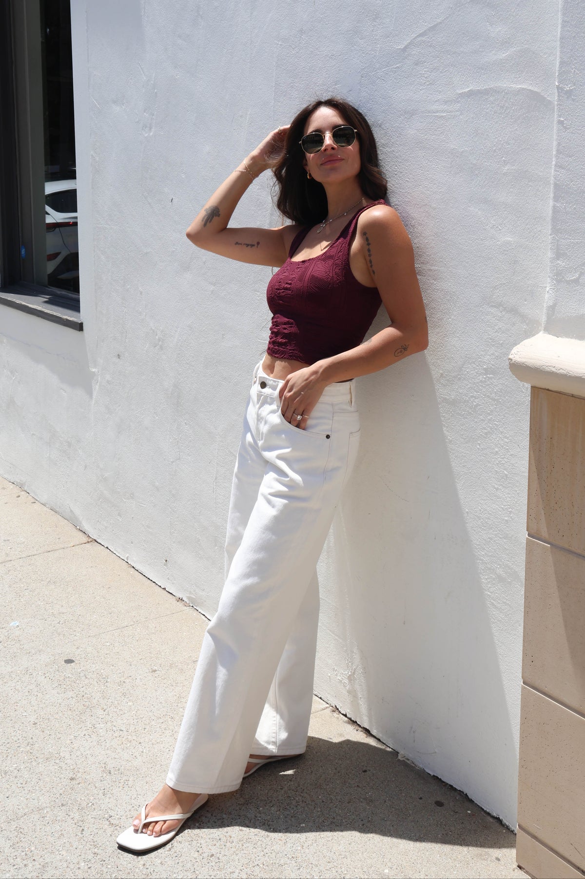 A woman stands against a white stucco wall wearing a sleeveless burgundy tank top, white wide-leg jeans, and white flat sandals.