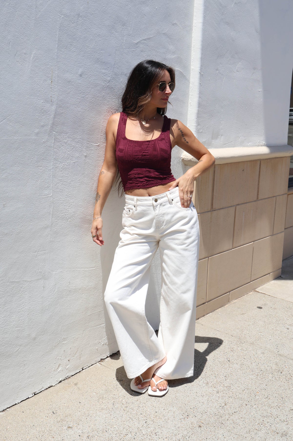 A woman posing outdoors against a white stucco wall, wearing a sleeveless burgundy crop top, white wide-leg pants, and white sandals.