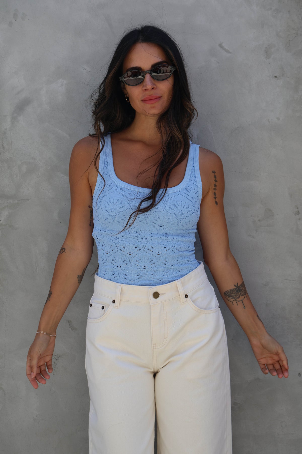 Woman wearing a blue tank top and white pants against a gray wall
