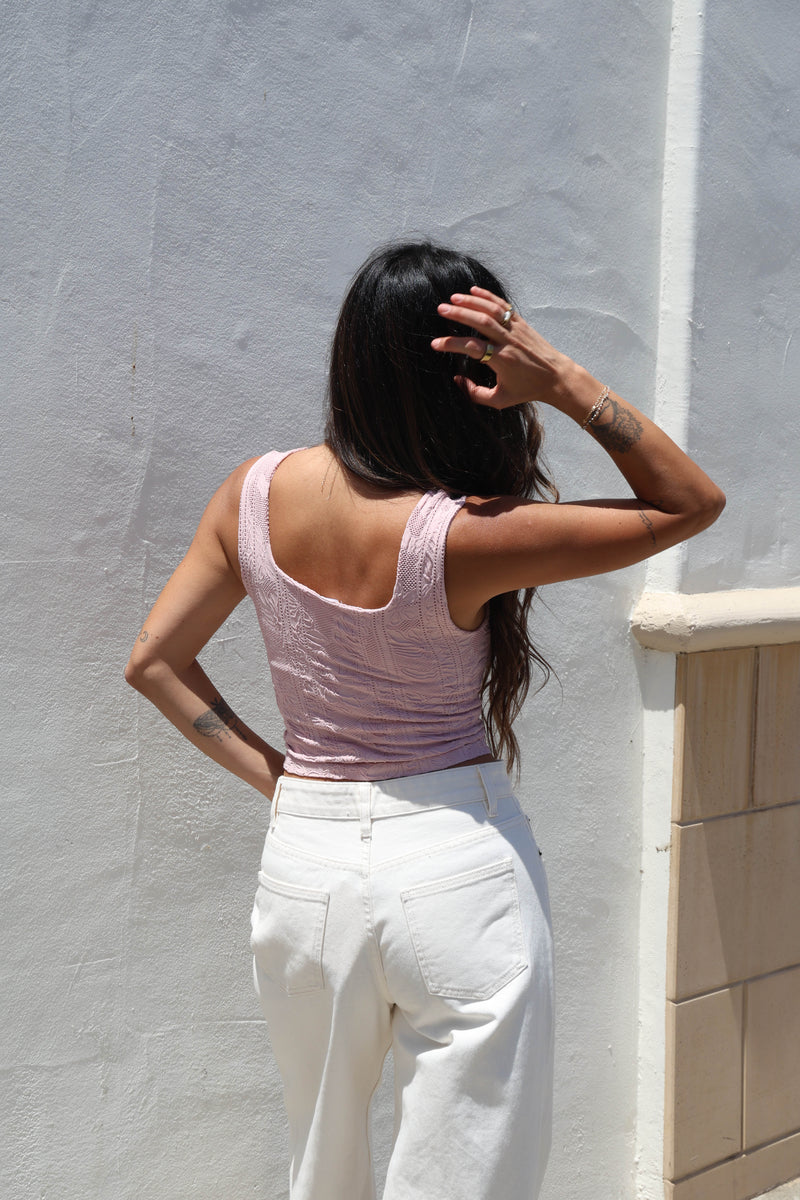 A woman wearing a light pink ribbed tank top and white jeans, standing outdoors against a white wall.
