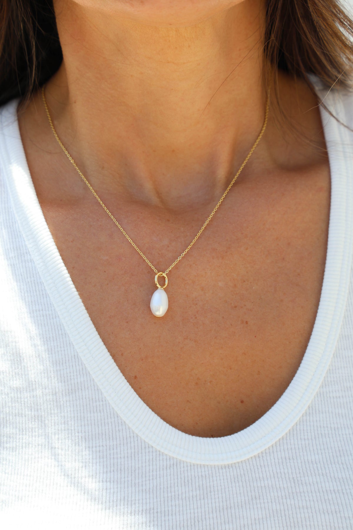 A close-up of a model wearing a delicate gold chain necklace with a teardrop-shaped white pearl pendant, styled with a white ribbed V-neck top.