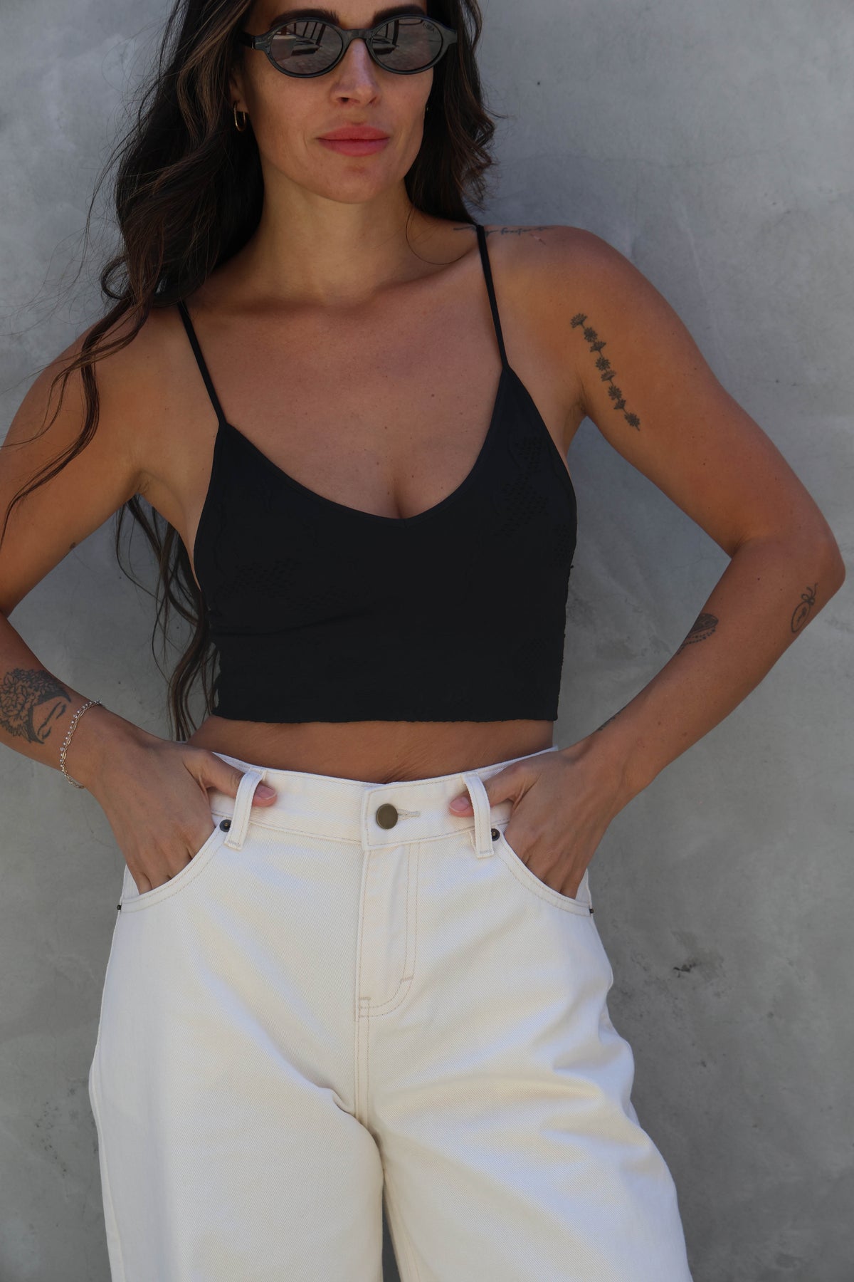 Woman wearing a black crop top and white pants against a gray wall.
