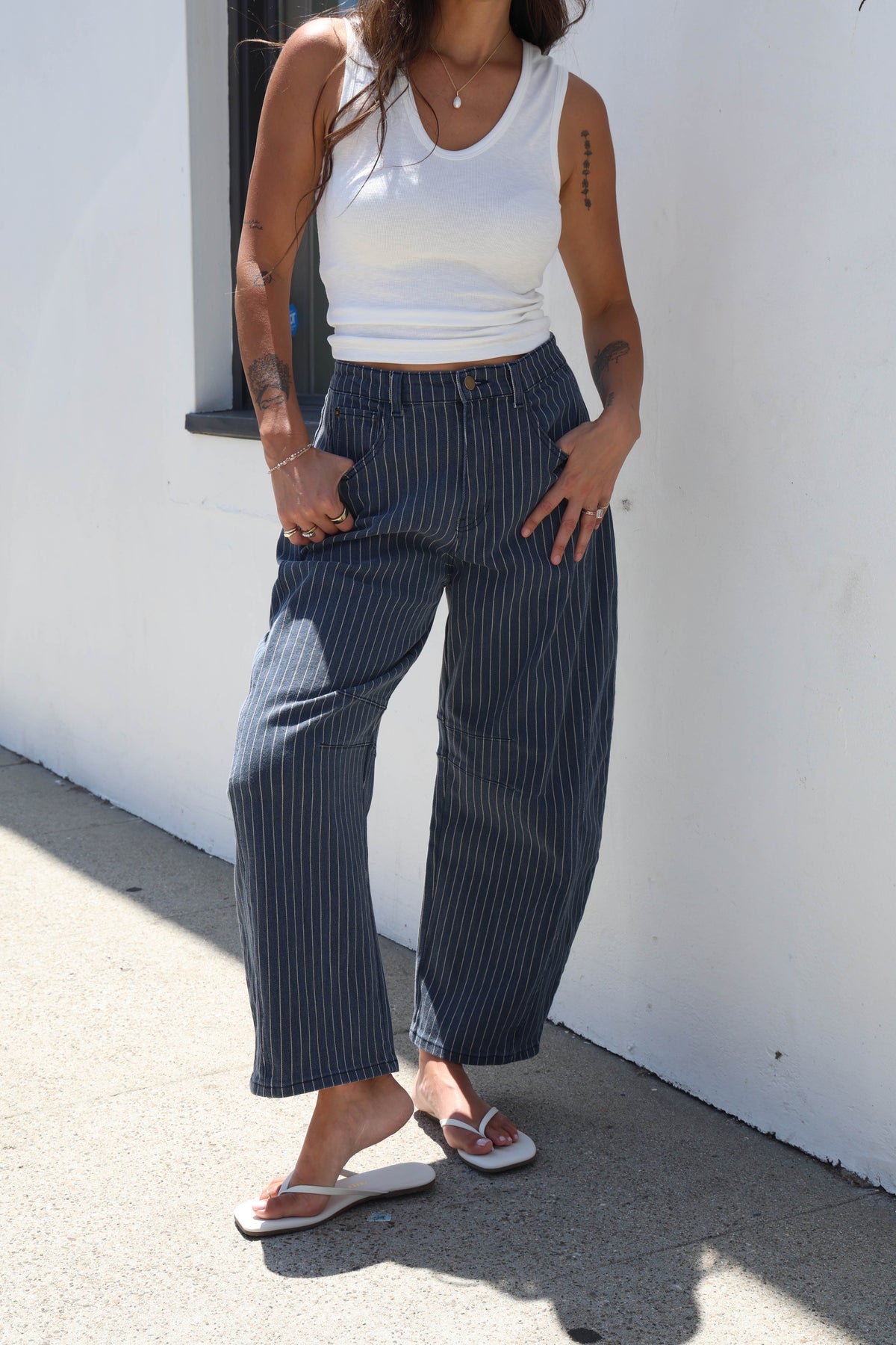 A woman wearing navy blue pinstriped wide-leg cropped pants, a white ribbed tank top, and white slide sandals, standing against a white stucco wall.
