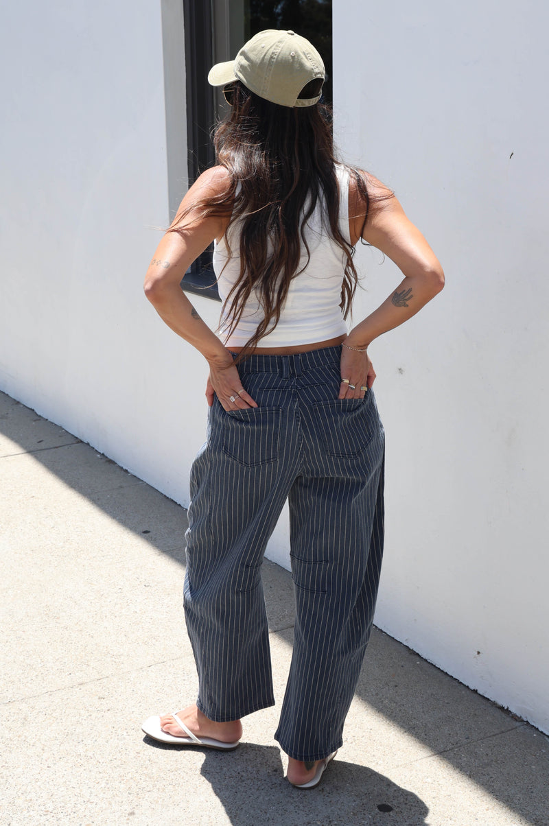 A woman stands outdoors against a white wall wearing high-waisted, wide-leg pinstripe trousers in navy and white, a white ribbed tank top, and a beige baseball cap.
