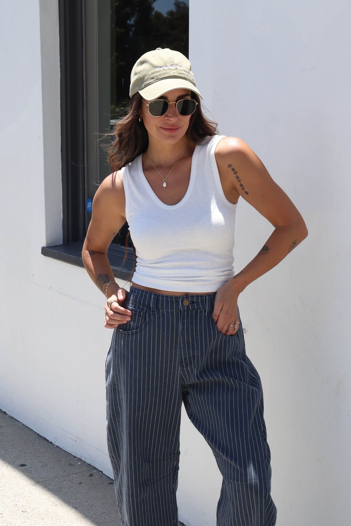 A woman posing outdoors in a white ribbed tank top, navy blue pinstriped wide-leg jeans, a beige baseball cap, and sunglasses against a white wall.
