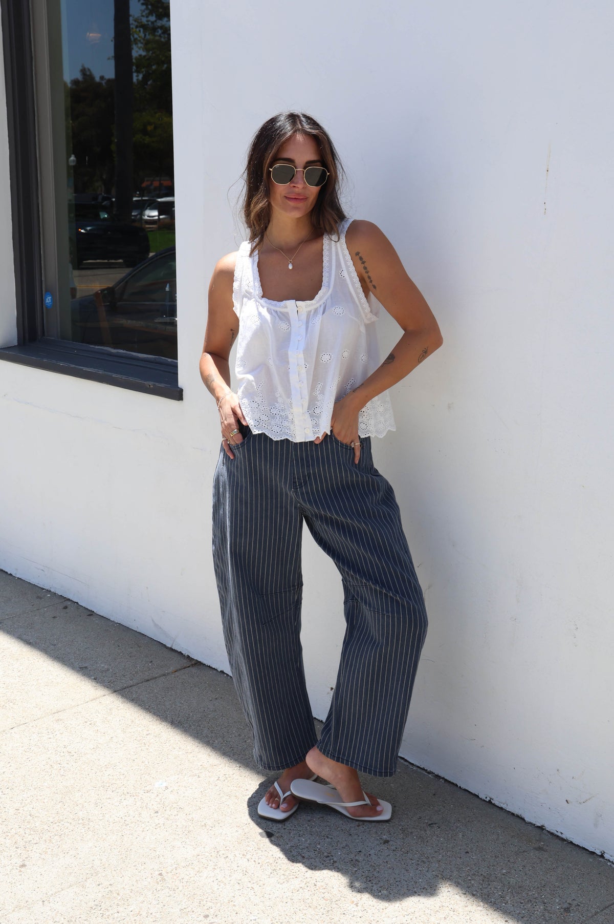 A woman wearing a white sleeveless ruffled top and dark gray pinstriped wide-leg pants stands against a white wall outdoors.