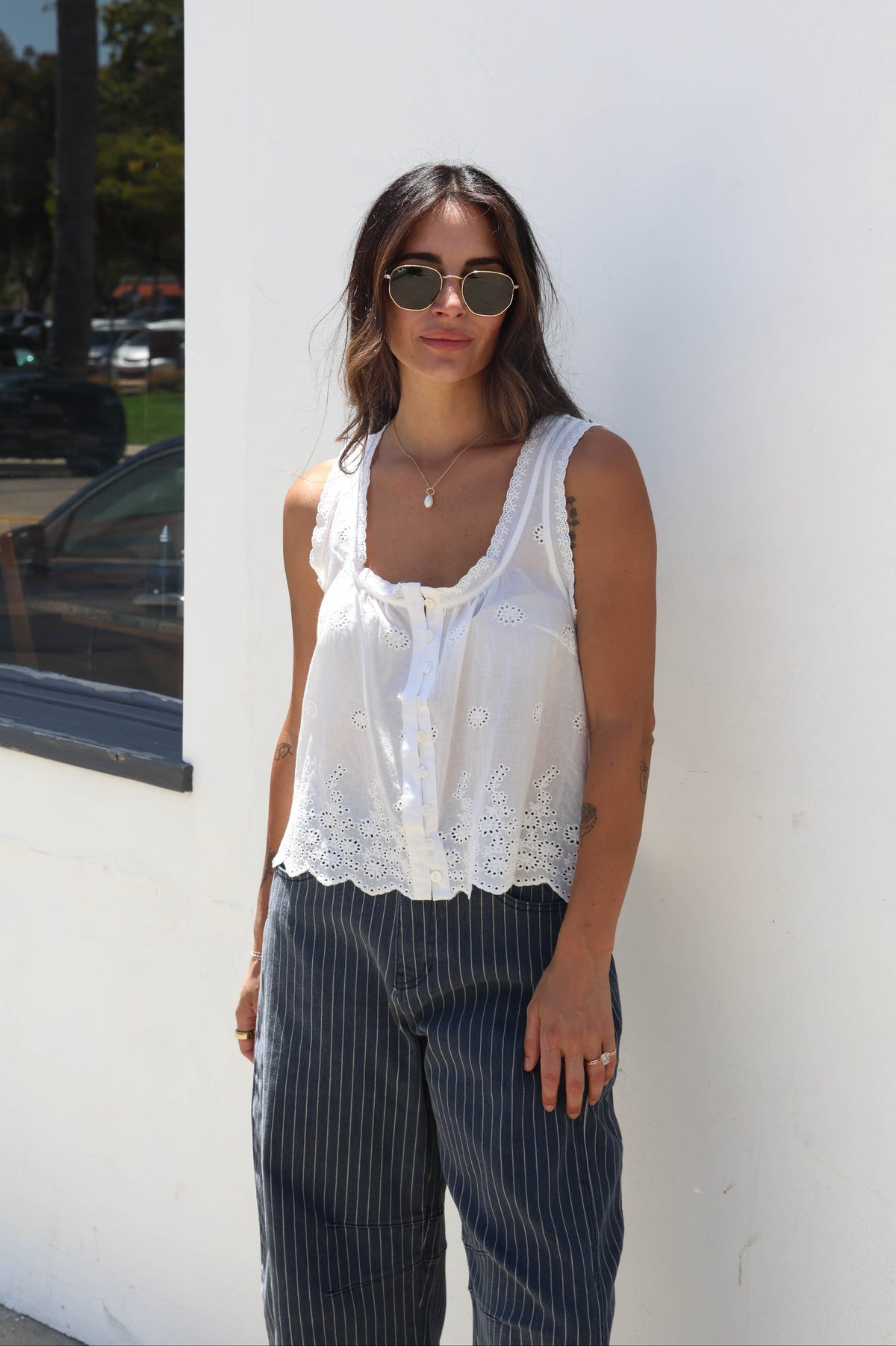 A woman wearing a white sleeveless lace-trim tank top and dark pinstriped pants, standing against a white wall outdoors.