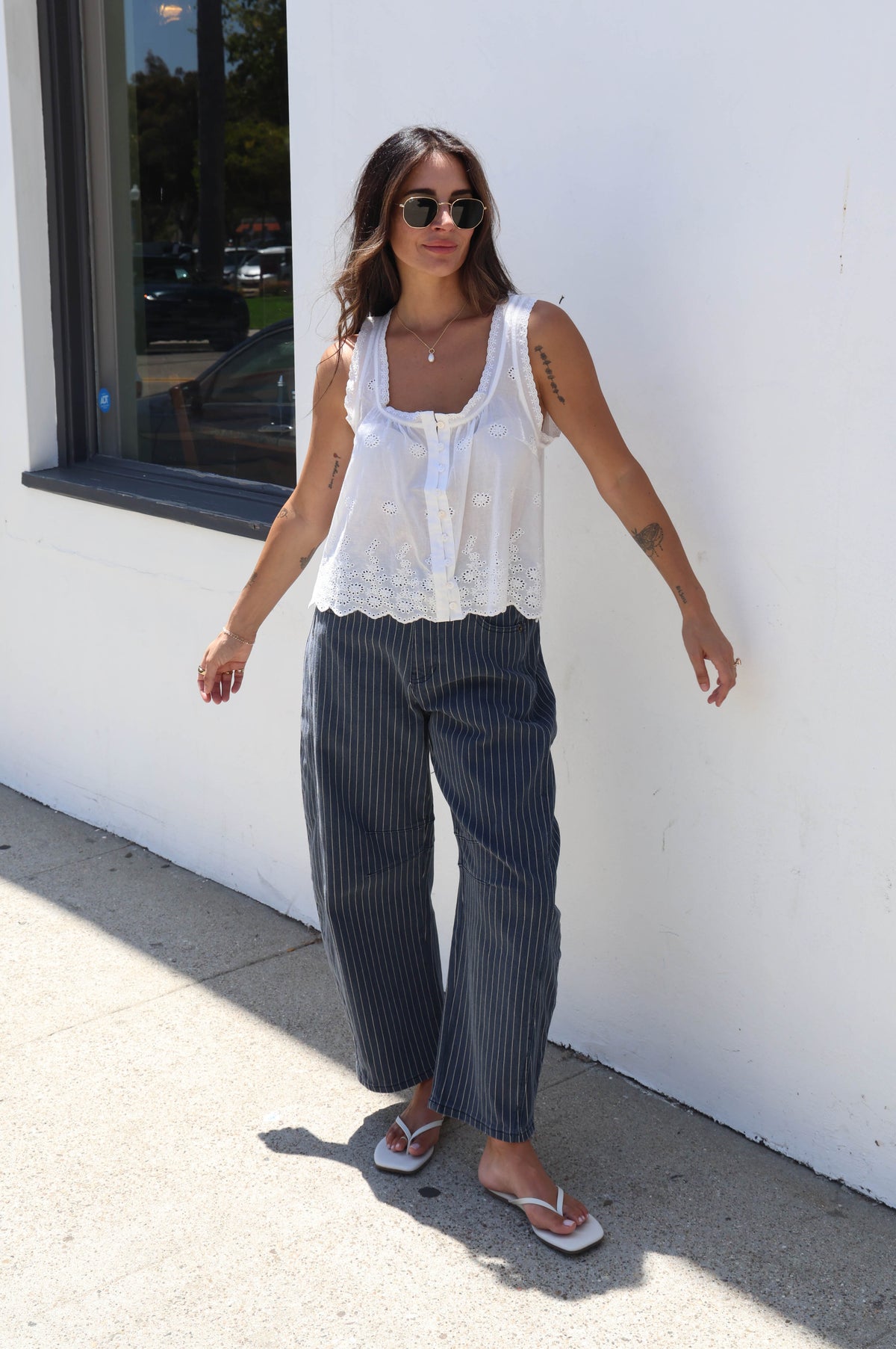 A woman stands outdoors against a white wall wearing a white sleeveless lace-trim top and dark pinstriped wide-leg pants, with flip-flop sandals and sunglasses.