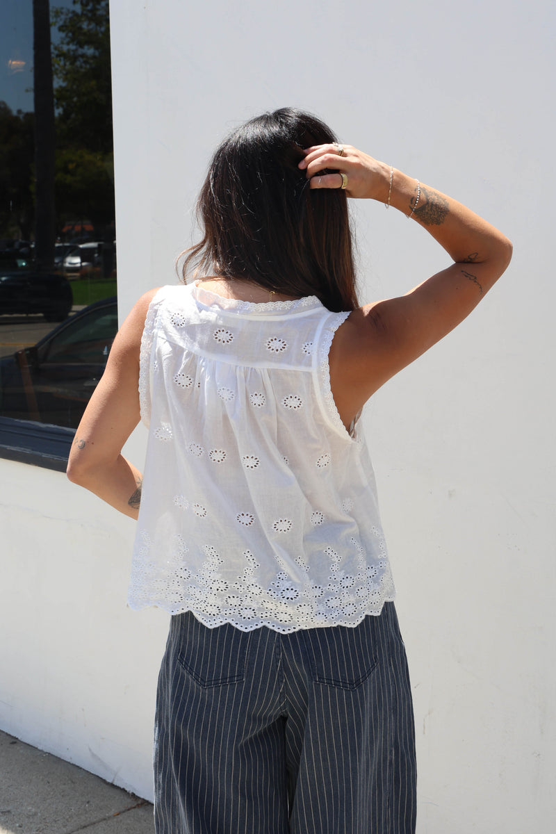 A woman shown from behind wearing a white sleeveless eyelet lace top and dark pinstriped pants, standing against a white wall.