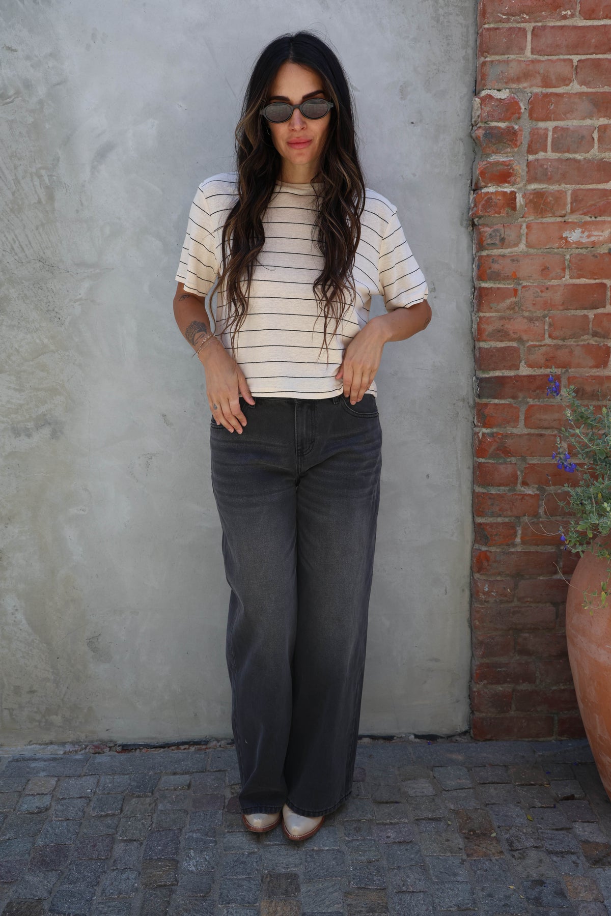 Woman wearing a striped shirt and dark jeans standing against a textured wall.