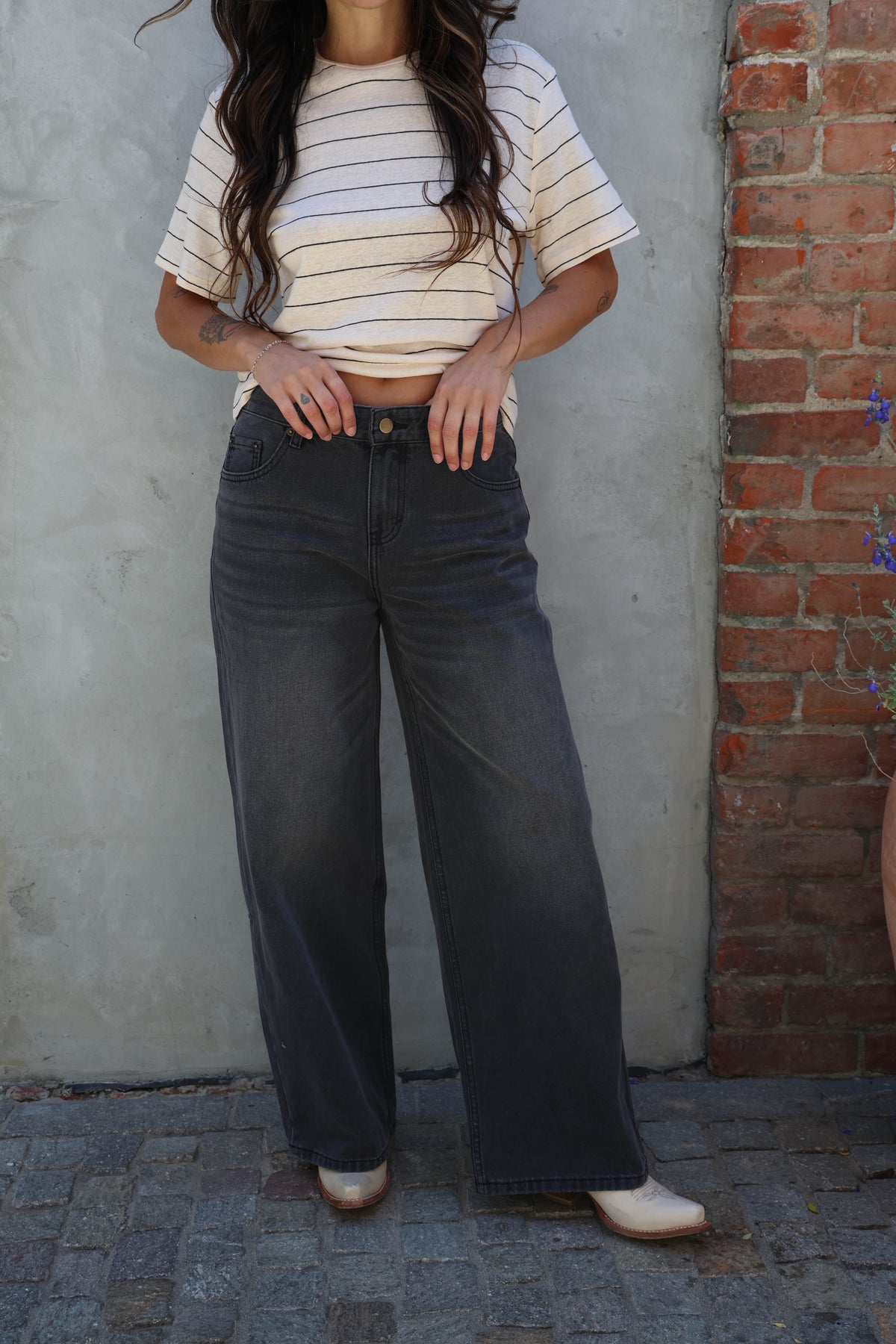 Person wearing a striped shirt and dark jeans standing against a gray wall with a brick column.