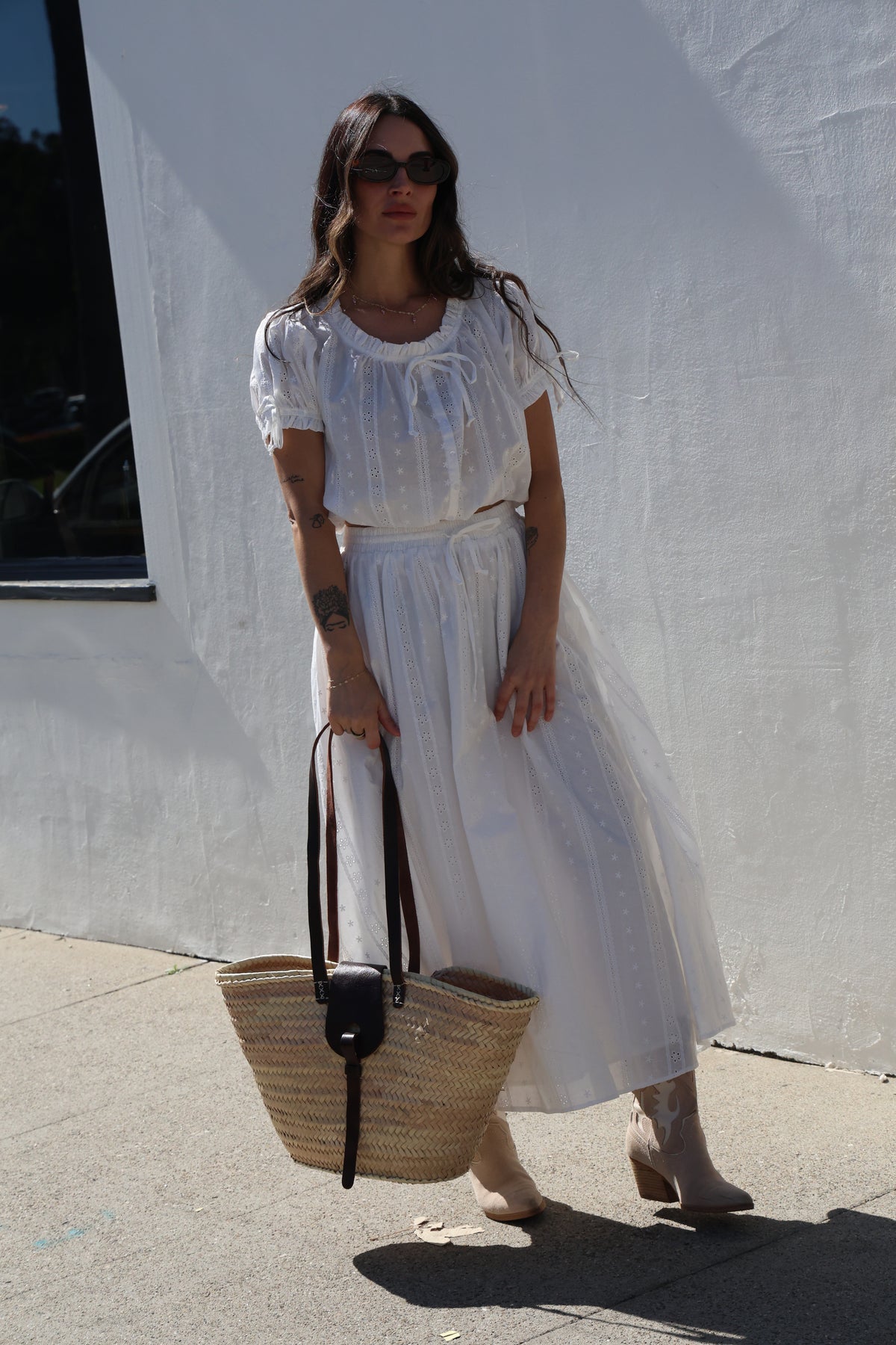 Woman in a white dress holding a woven bag on a sidewalk.