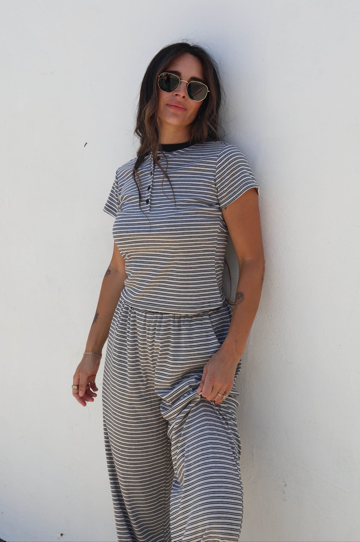 A woman wearing a gray and white striped short-sleeve jumpsuit and sunglasses, posing against a plain white wall.