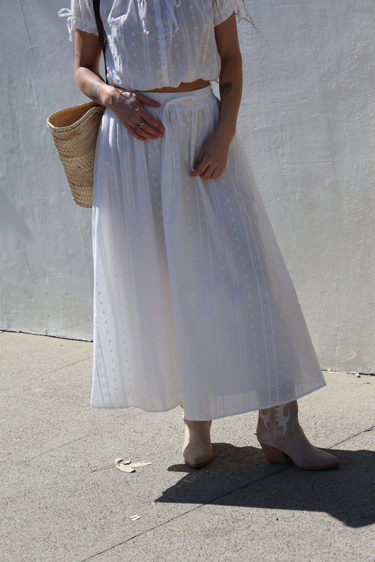 Person wearing a long white dress holding a straw bag on a sidewalk.