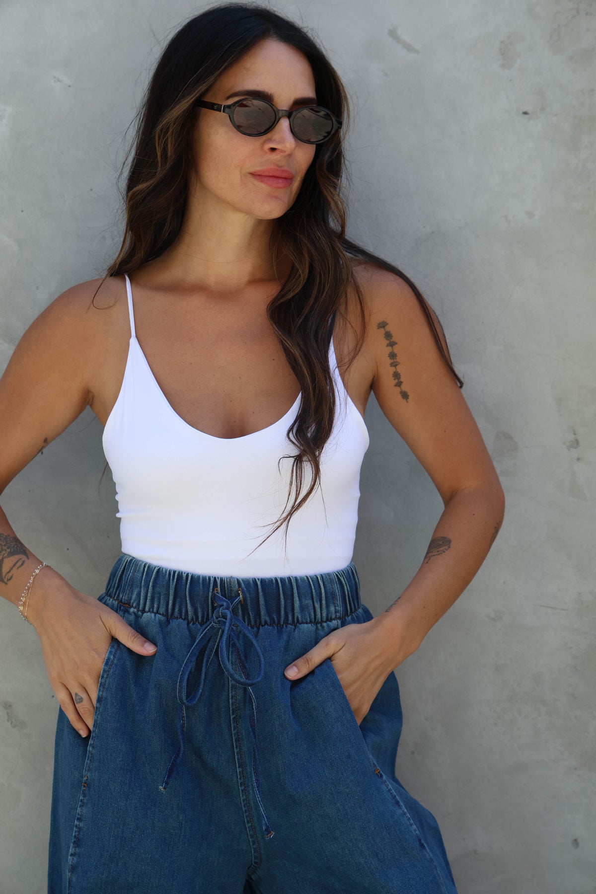 Woman wearing a white tank top and blue jeans against a gray wall