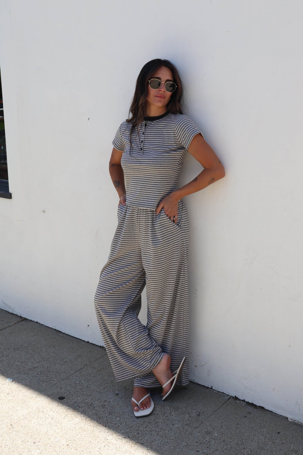 A woman wearing a beige and black striped short-sleeve jumpsuit with wide legs, standing against a white wall.